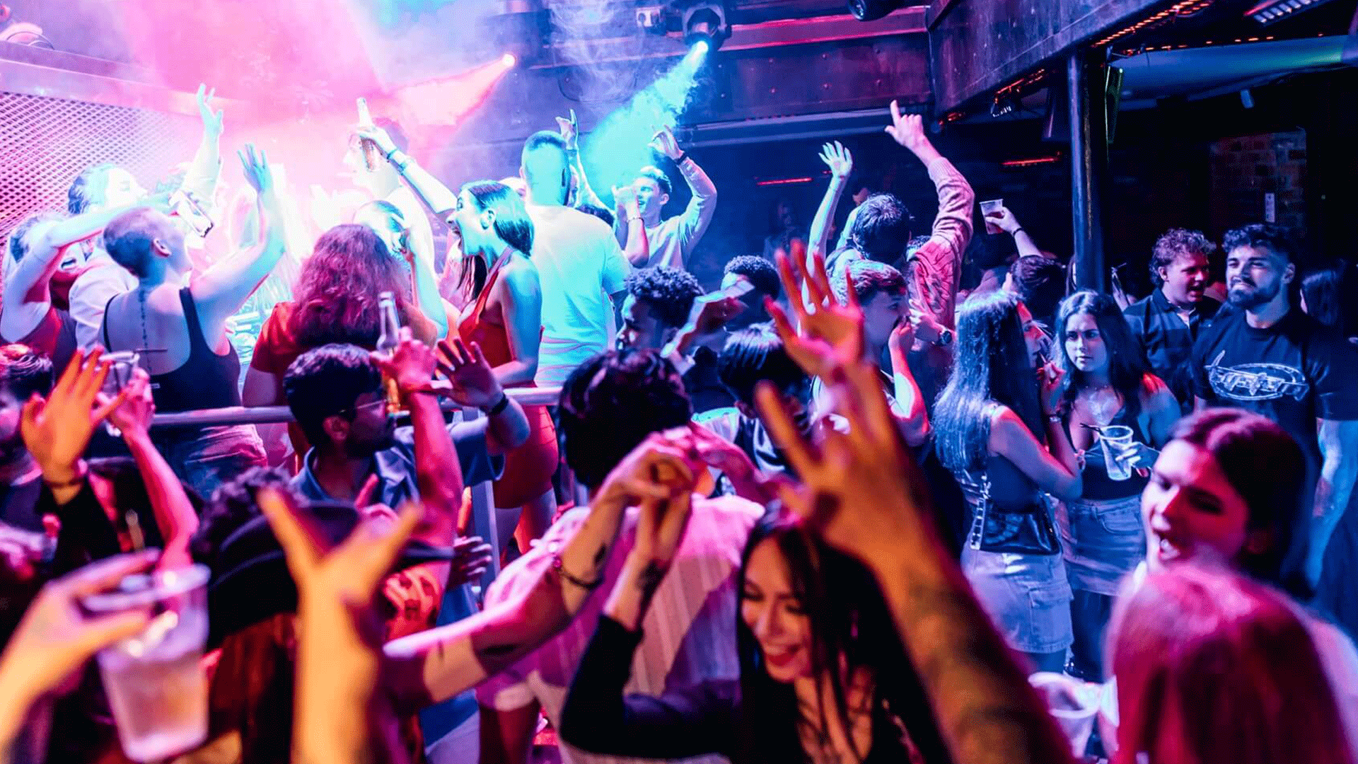A vibrant scene inside a nightclub with people dancing energetically under colorful lights. Many attendees are raising their hands and enjoying the music. This represents a lively club night atmosphere at Thompsons Garage in Belfast, showcasing the nightlife and house music culture.