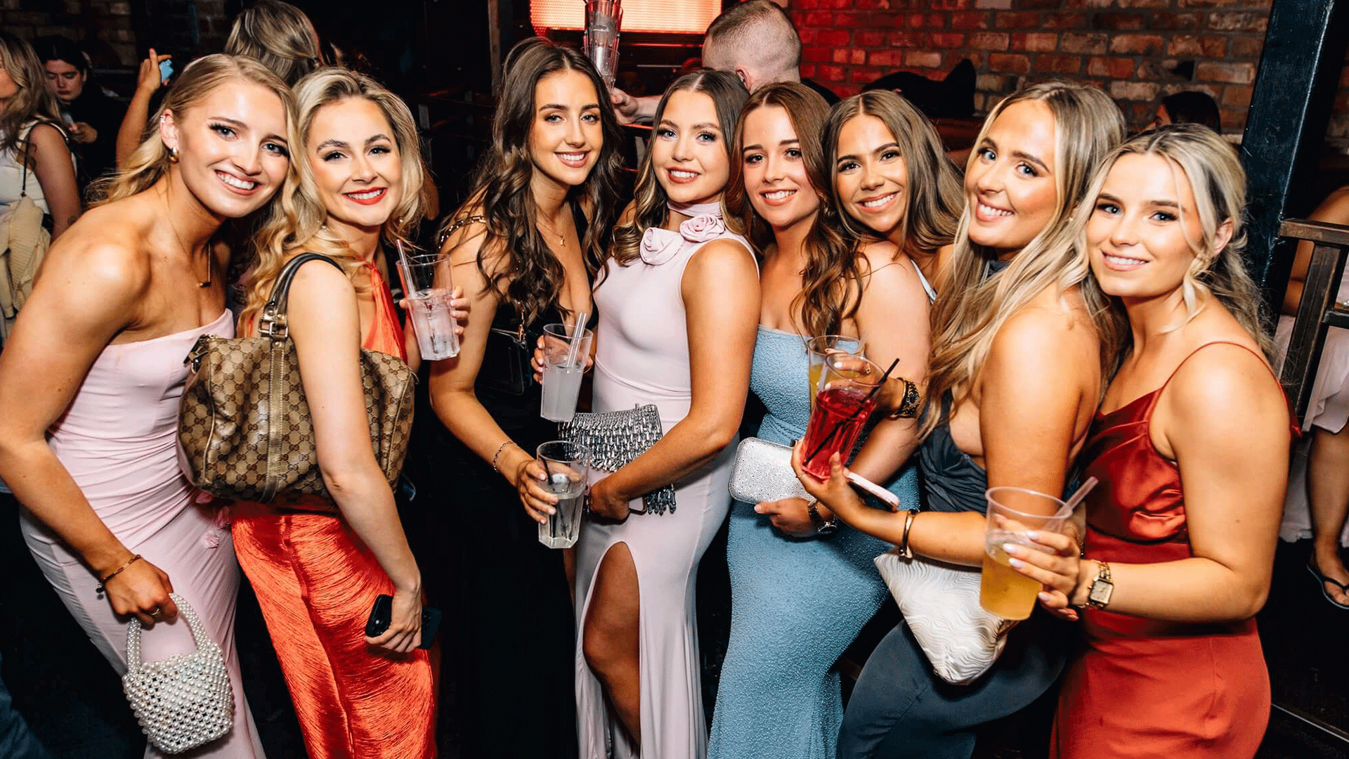 A group of eight women posing together in a vibrant nightclub setting. They are dressed in stylish evening attire and holding drinks, smiling for the camera. The scene captures the lively atmosphere of Belfast nightlife at Thompsons Garage during a club night featuring house music.