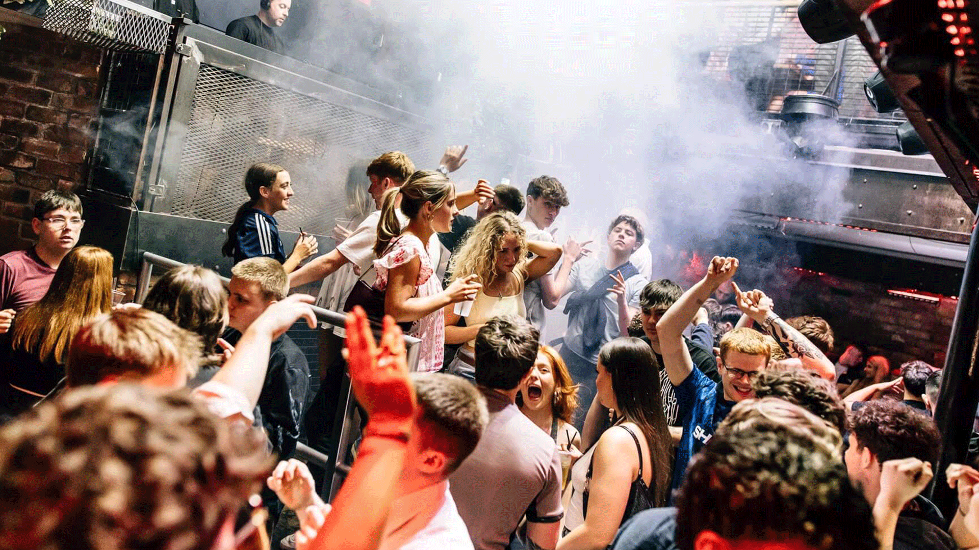 A crowded nightclub scene with young people dancing under colorful lights and a light haze. The atmosphere is lively, showcasing the vibrant nightlife of Belfast, particularly at Thompsons Garage during a house music DJ event.
