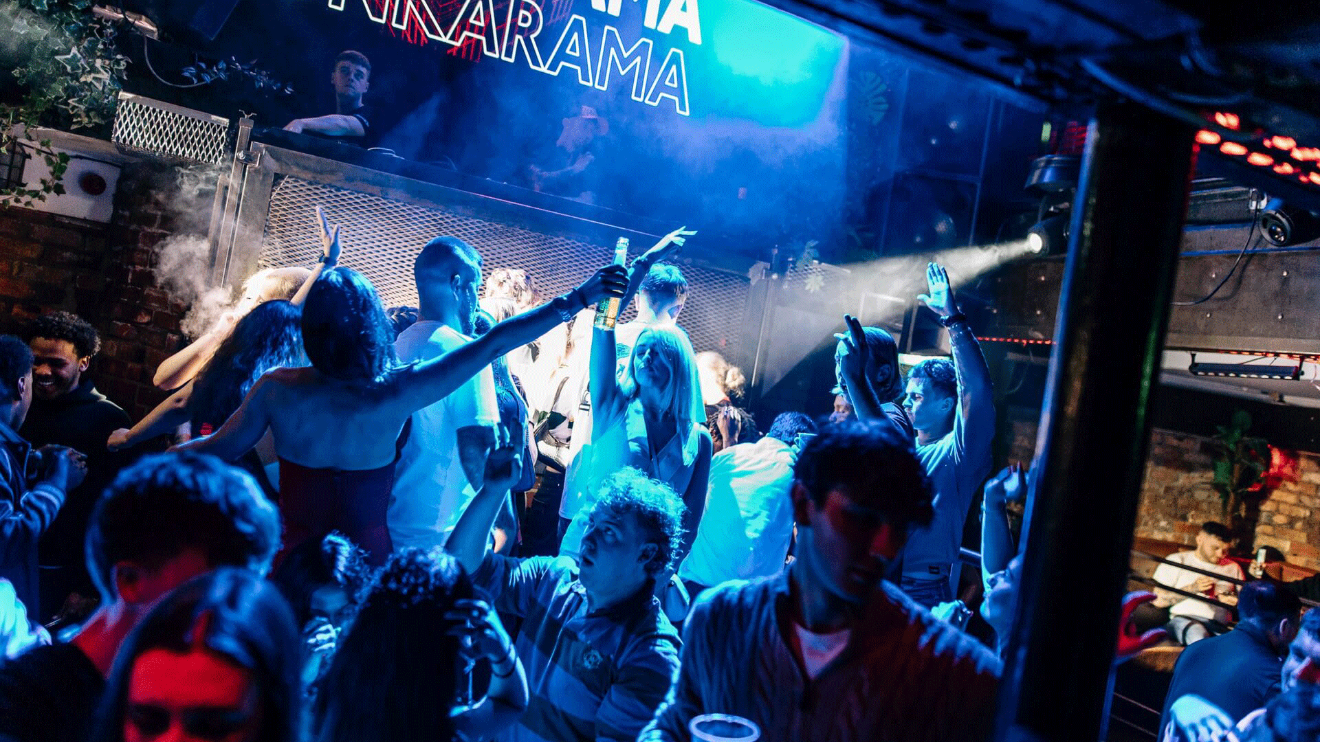A crowded nightclub scene in Belfast with people dancing and enjoying a DJ event. The venue features vibrant lighting, and the atmosphere suggests lively house music. The Thompsons Garage sign is visible, highlighting Belfast nightlife.