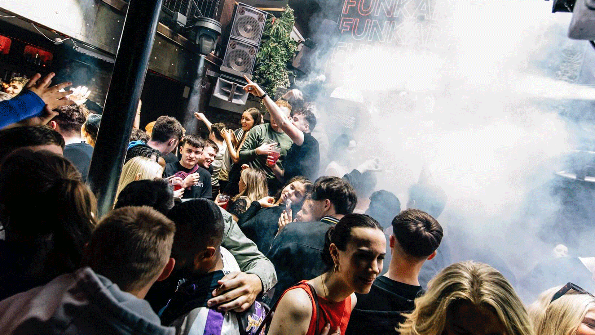 A crowded nightclub scene with people dancing and enjoying drinks amidst colorful lights and smoke. The atmosphere captures the vibrancy of Belfast nightlife during a DJ event featuring house music at Thompsons Garage.