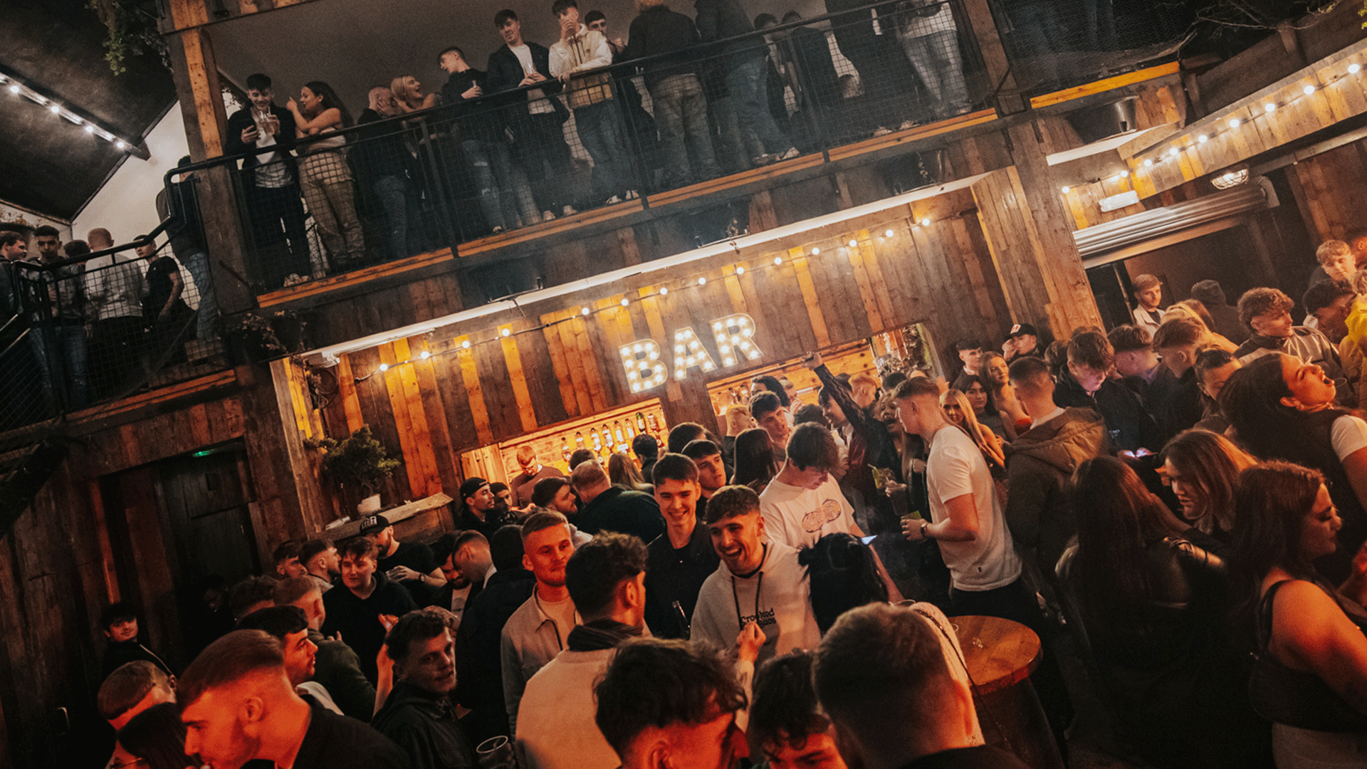 A lively crowd socializing in a wooden-themed interior of a nightclub, with people mingling near a bar illuminated by lights. The scene captures the energetic atmosphere of Belfast nightlife at Thompsons Garage during a club night featuring house music and DJ events.