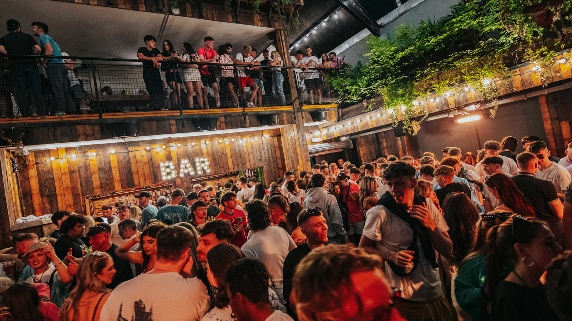 A lively scene at Thompsons Garage, a Belfast Nightclub, filled with people enjoying a club night, dancing, and socializing. The venue has a bar prominently displayed, decorated with string lights and plants.