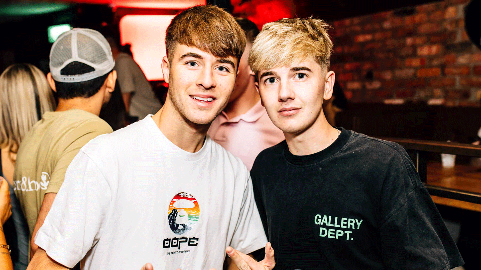 Two young men posing for a photo in a nightlife setting, with one wearing a white shirt featuring a colorful graphic and the other in a black shirt with "GALLERY DEPT." text. The background shows a lively atmosphere typical of a club night.