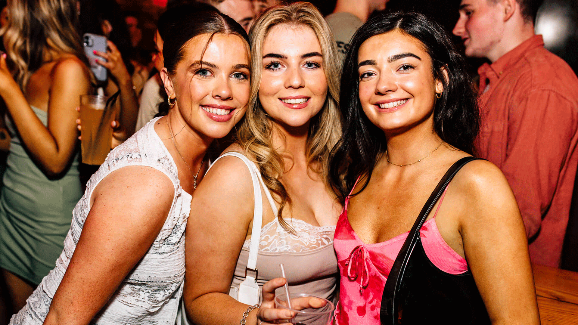 Three young women posing together at a Belfast nightclub during a lively club night. The interior is dimly lit, and a DJ can be seen in the background, contributing to the vibrant Belfast nightlife. They are enjoying the atmosphere.
