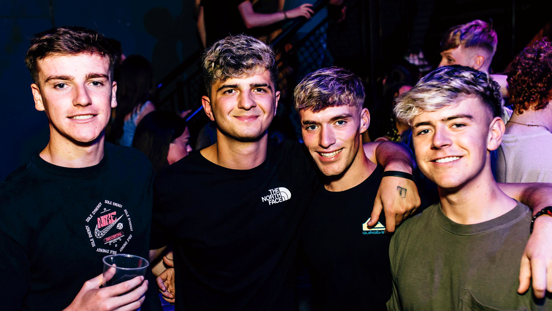 Four young men smiling together at a lively club scene, dressed casually. The background features a blurred crowd, suggesting an energetic atmosphere typical of a Belfast Nightclub like Thompsons Garage during a Club Night with House Music.
