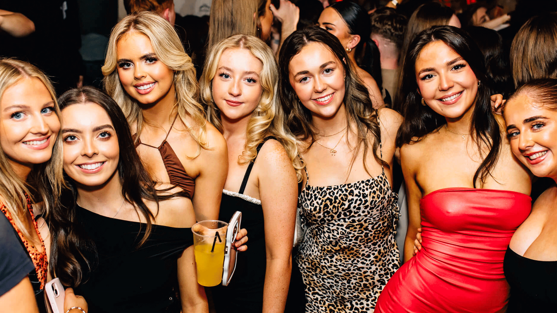 A group of six women posing together in a vibrant nightlife setting, dressed in stylish outfits. The background features a lively crowd, indicative of a club night atmosphere, possibly at a Belfast nightclub.