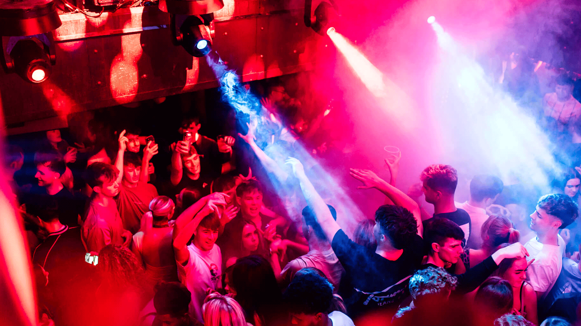 A crowded nightclub scene at Thompsons Garage in Belfast, filled with energetic people dancing under colorful lights and fog. The image captures the vibrant atmosphere of a club night featuring house music and a DJ event.