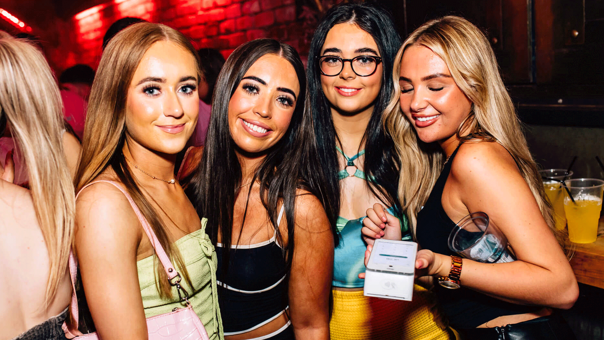 Four young women smiling and posing together in a lively setting, with dim lighting and colorful backgrounds, indicative of a vibrant Belfast Nightclub atmosphere, possibly Thompsons Garage, reflecting Belfast Nightlife and Club Night vibes.