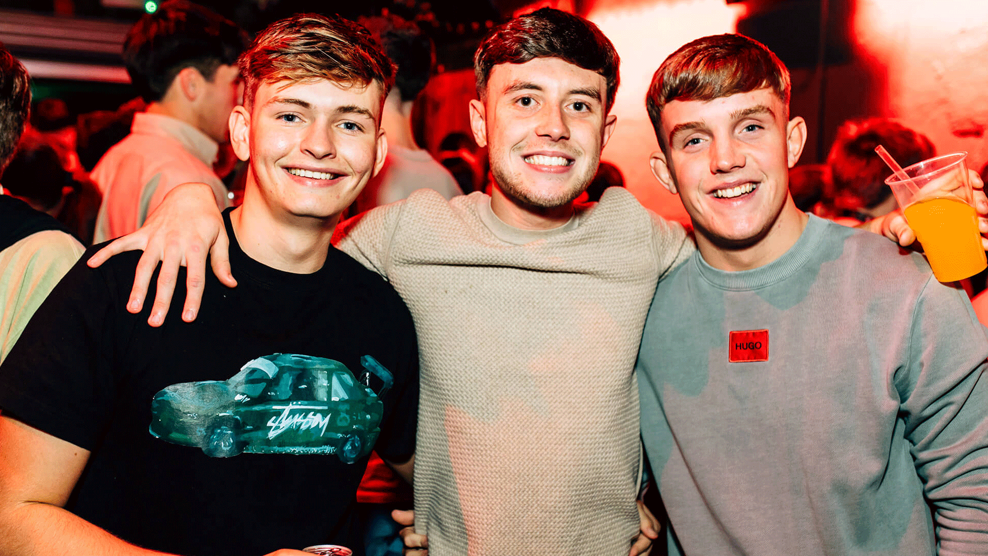 Three young men smiling and posing for a photo in a nightlife setting, likely at Thompsons Garage in Belfast. The background features a vibrant atmosphere, indicative of Belfast nightlife with club lights.