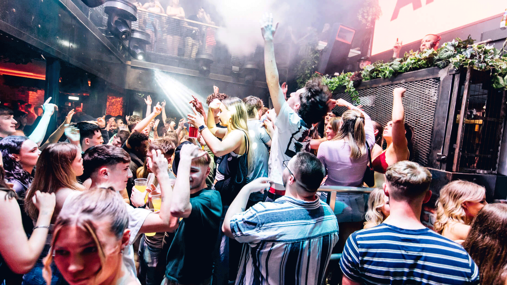 A lively scene inside a Belfast nightclub packed with people dancing. Attendees enjoying a DJ event with raised hands amidst colorful lights and smoke, embodying the vibrant Belfast nightlife, particularly at Thompsons Garage.