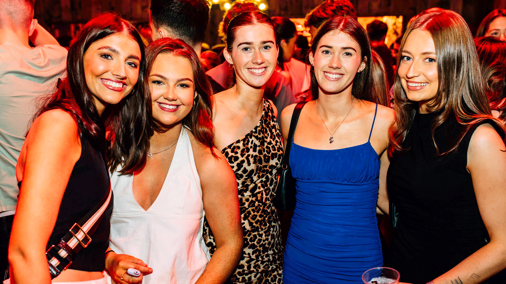 Five young women posing together in a vibrant setting, smiling at the camera. The background is filled with club lights, indicative of a lively atmosphere likely at a Belfast Nightclub, possibly Thompsons Garage during a Club Night featuring House Music and a DJ Event.
