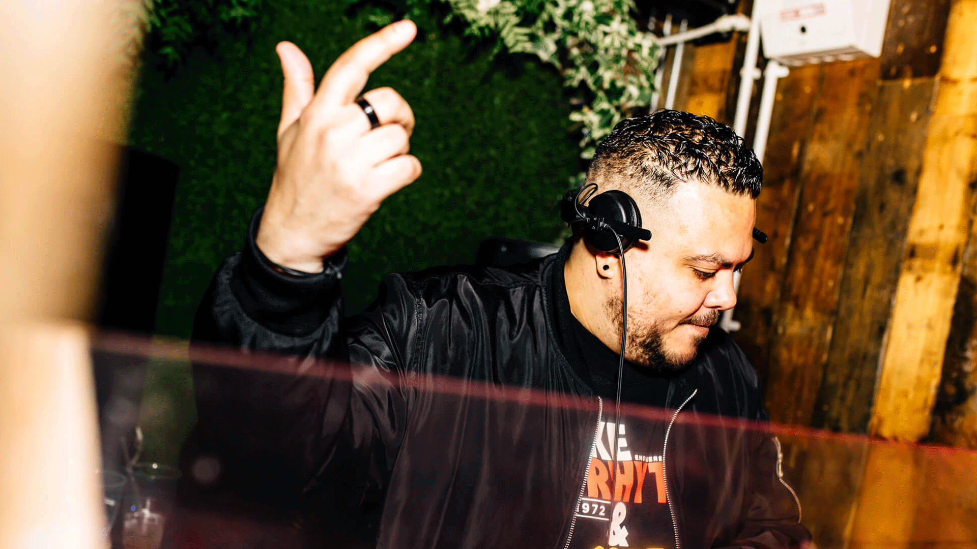 A DJ performing at a vibrant nightclub, wearing a black jacket, with headphones on and one hand raised, set against a backdrop of greenery and wooden decor, highlighting the Belfast Nightlife and House Music scene.