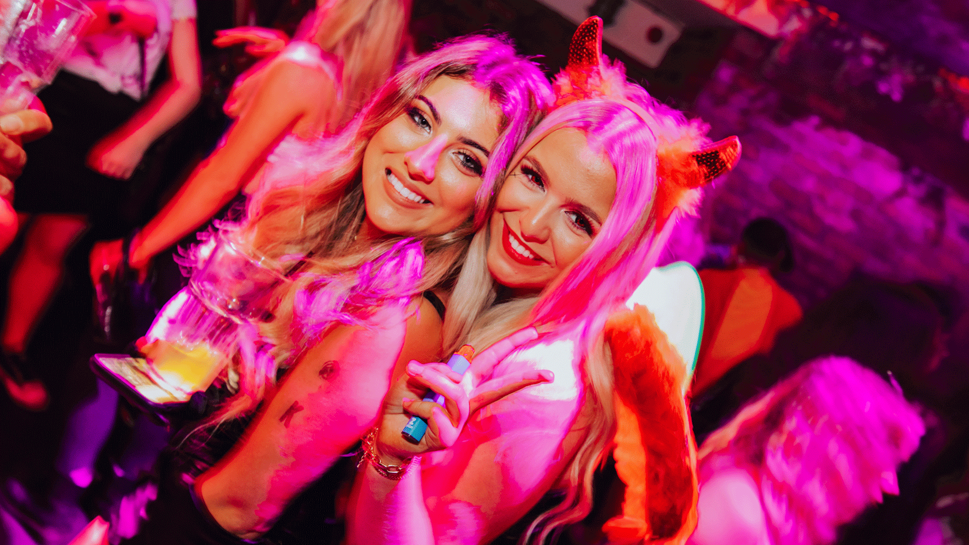 Two women posing for a photo at a nightclub, wearing casual outfits. One woman has long blonde hair and smiles with drinks in hand, while the other sports devil horns and a playful expression. The setting reflects the vibrant atmosphere of a Belfast nightclub.
