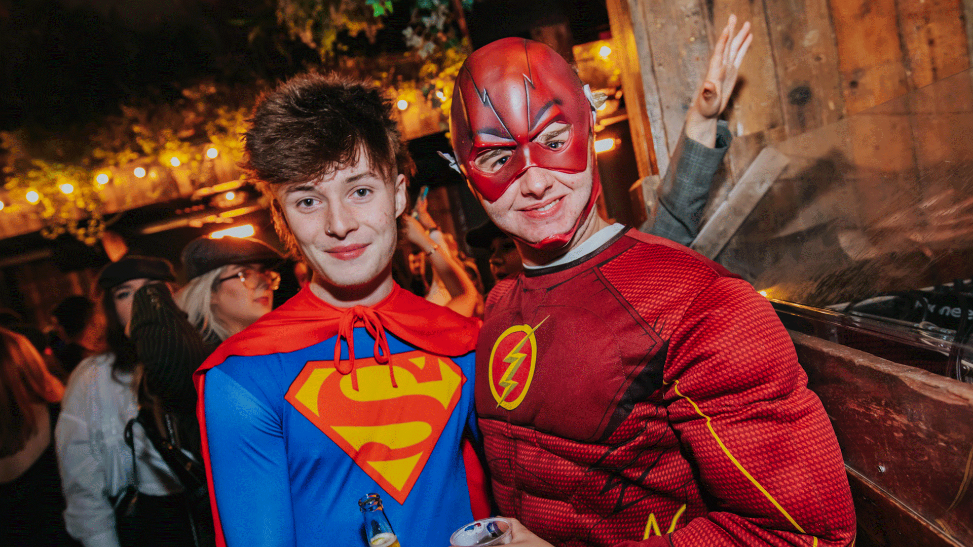 Two men in superhero costumes at a lively event in a venue with warm lighting. One is dressed as Superman with a red cape, and the other as The Flash in a red and yellow suit, both enjoying the atmosphere of a Club Night.