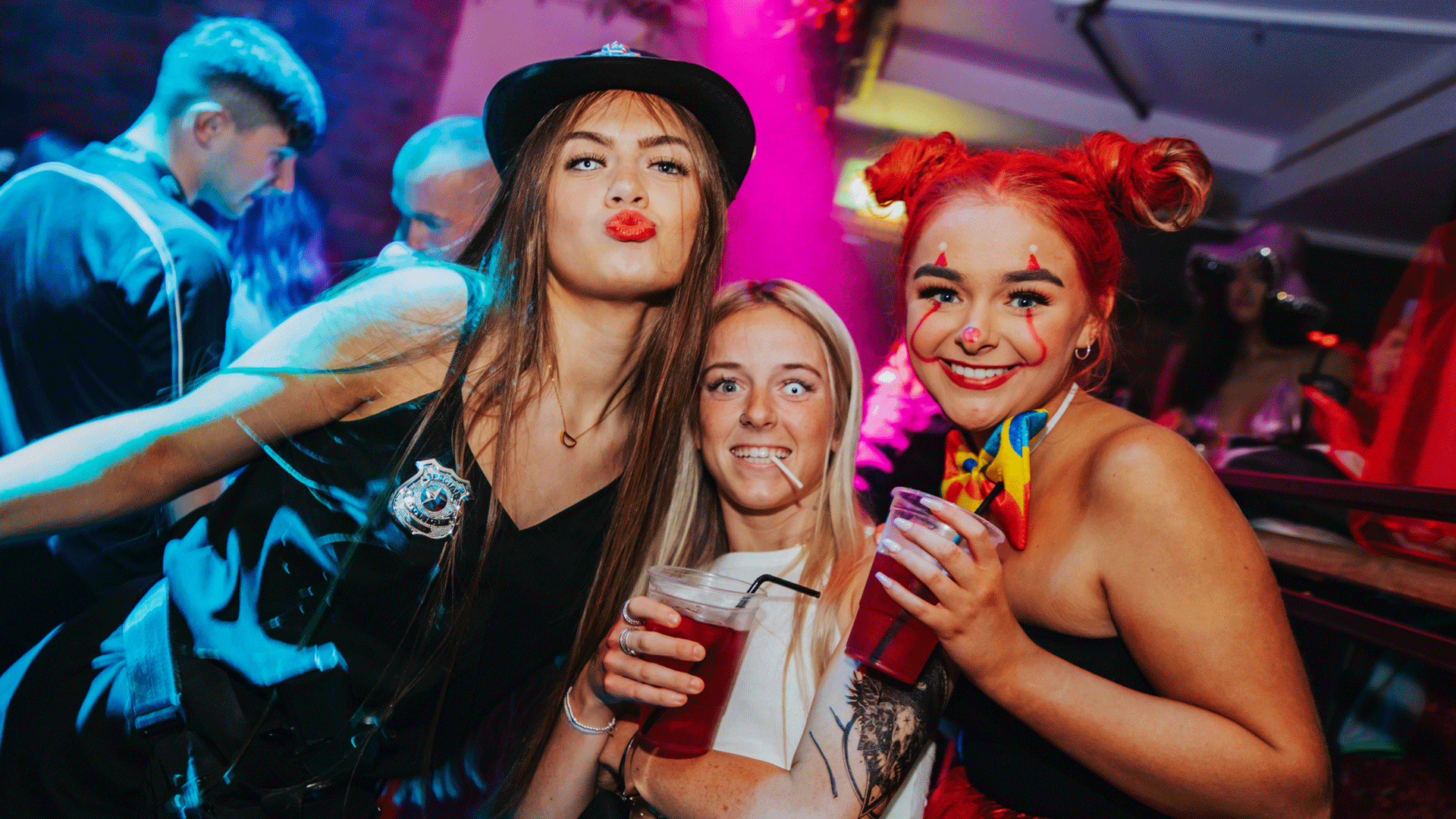 Three women posing together at a vibrant Belfast Nightclub during a club night. They are dressed in playful outfits, holding drinks, with lively party lights in the background. The image captures the energetic atmosphere of Belfast nightlife, highlighting a house music DJ event.