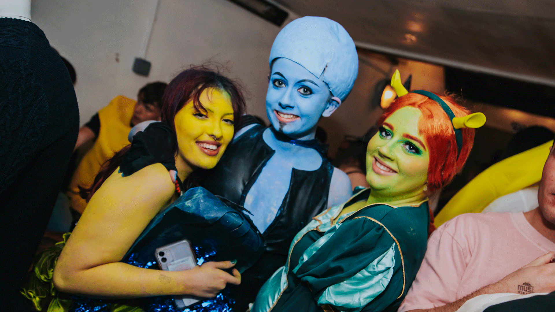 Three friends posing together at a themed event, wearing creative costumes with bright makeup. One has blue skin and an extraterrestrial look, while the others have vibrant green and yellow hues. The background hints at a festive atmosphere, likely part of a Club Night in Belfast.