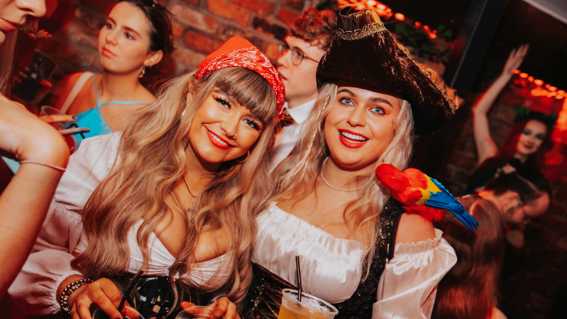 Two women posing together at a party, wearing costumes. One woman has a red bandana and a drink, while the other wears a pirate outfit with a colorful parrot on her shoulder. The background features dim lighting and a brick wall, suggesting a lively atmosphere typical of a Belfast Nightclub event.