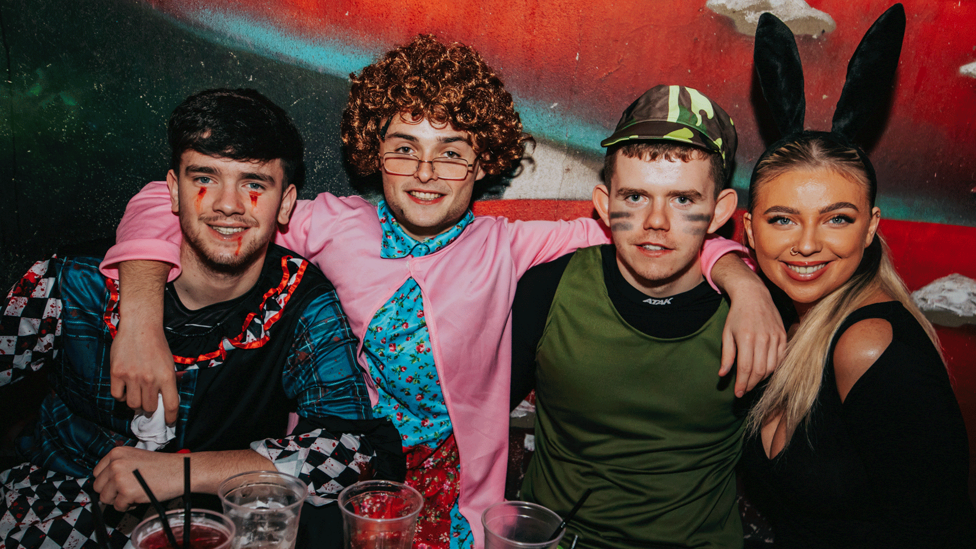 A group of four friends posing together in a vibrant setting at a Belfast nightclub. They are dressed in various costumes, enjoying a club night with drinks on the table. The background features colorful decorations, suggesting a lively atmosphere for a DJ event featuring house music.