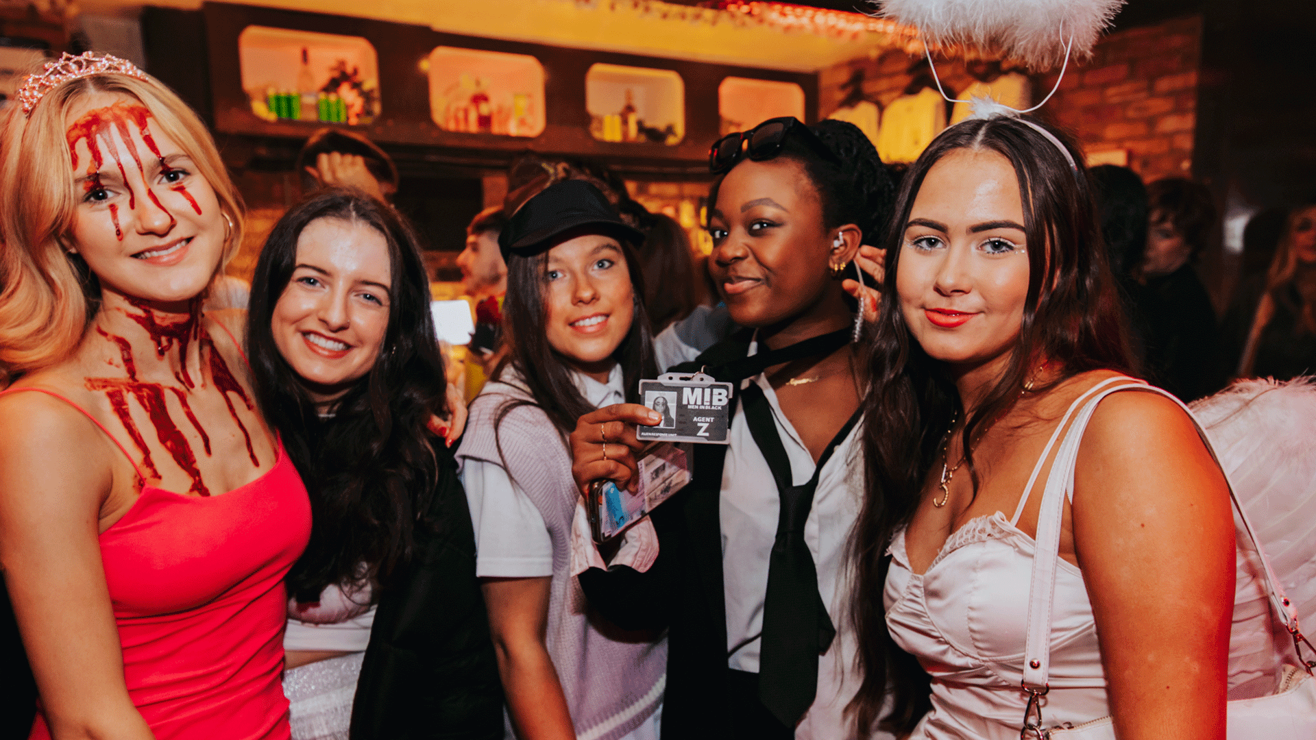 A group of five women posing together at a Belfast nightclub, dressed in various costumes for a themed club night event. The atmosphere is lively, reflecting Belfast nightlife, with Halloween-inspired attire present.