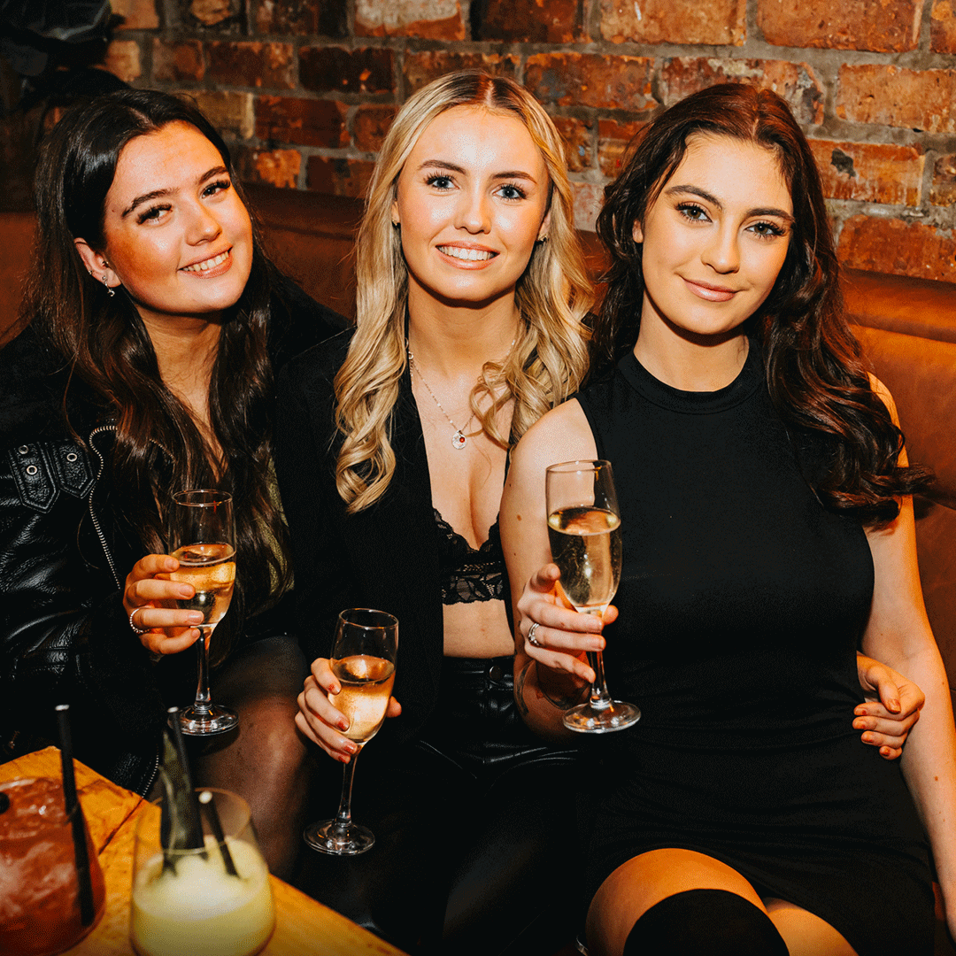 Three women sitting together in a cozy venue, each holding a glass of champagne. The background features exposed brick walls, creating a festive atmosphere. This image showcases a group enjoying a night out, relevant to Thompsons Belfast Night Club NightClub Staff Party Venue Hire.