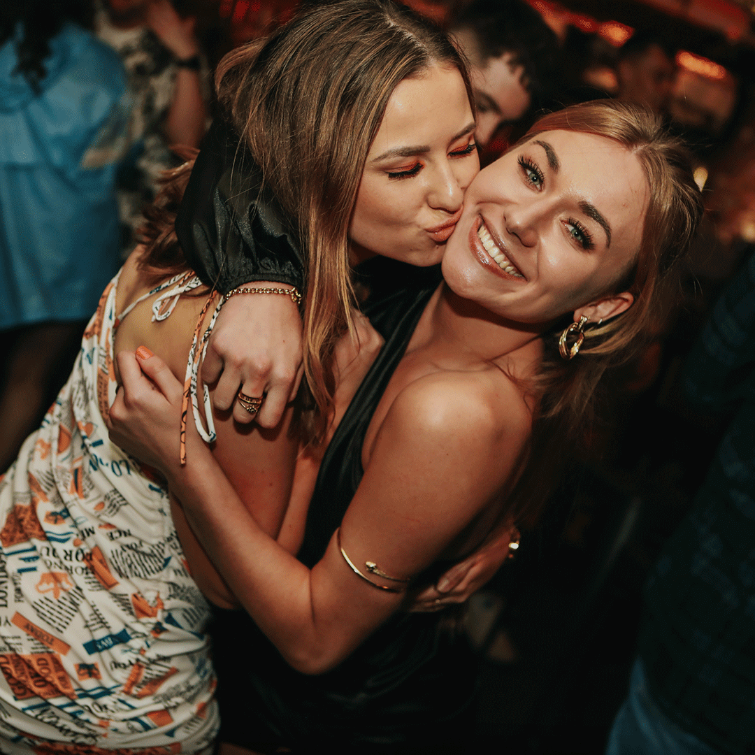 Two women smiling and embracing each other while at a vibrant party. The background is filled with blurred figures, suggesting a lively atmosphere. The image reflects a fun moment typical of nightlife events, relevant for Thompsons Belfast Night Club NightClub Staff Party Venue Hire.