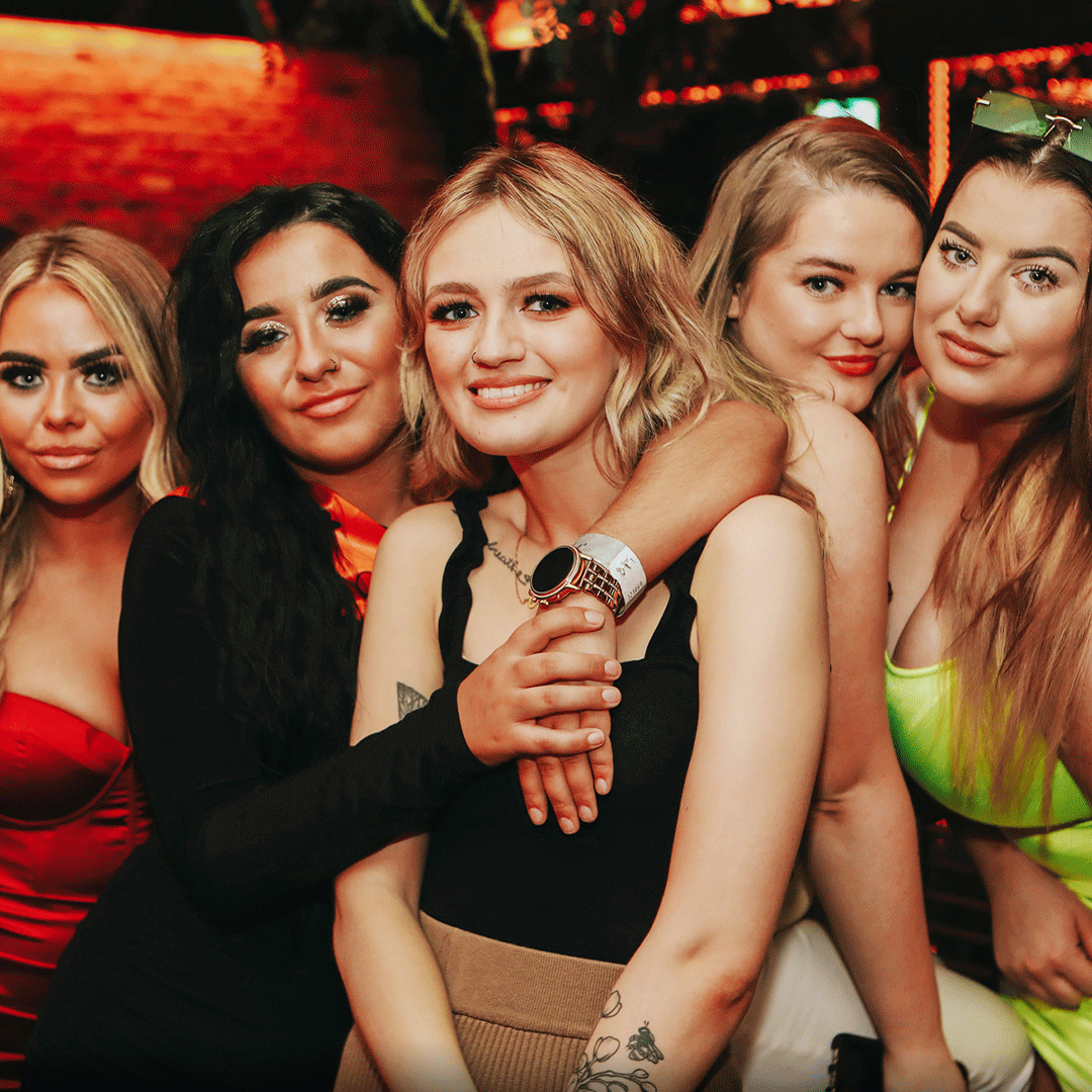 A group of five women posing together in a vibrant nightclub setting. They are smiling and dressed in stylish party outfits. The background features warm lighting and décor typical of a lively nightlife venue, ideal for a party or event.