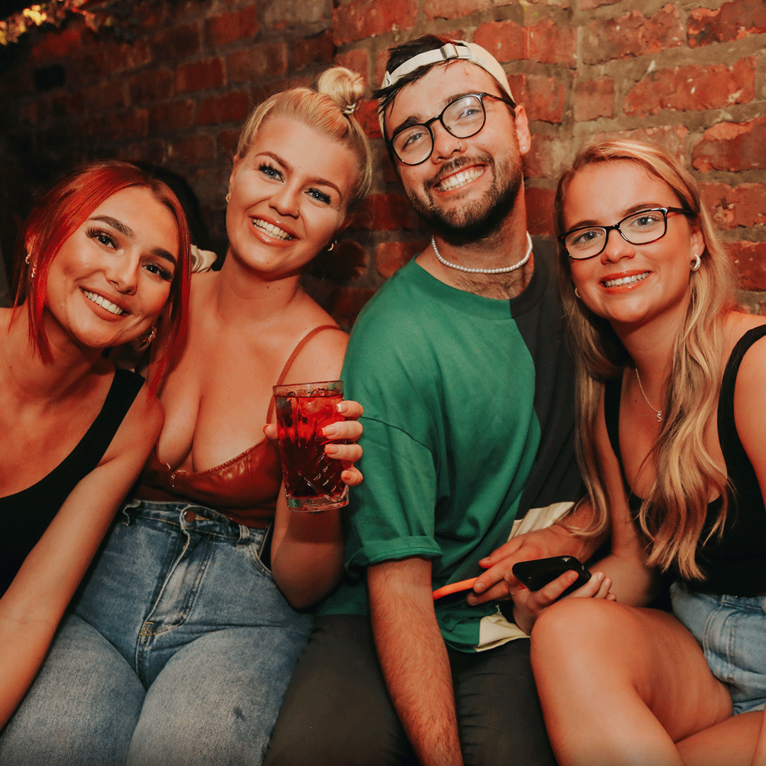 Four friends sitting together in an informal social setting, smiling and enjoying drinks. The group is illuminated by warm lighting in a cozy venue, showcasing a friendly and vibrant atmosphere, suitable for a night out at Thompsons Belfast Night Club.