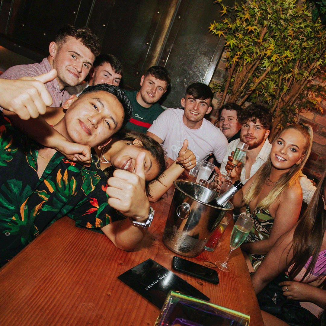 A group of young adults posing for a photo at a party in a lively venue. They are smiling and giving thumbs up, with drinks on the table, surrounded by greenery. This image reflects a celebratory atmosphere at Thompsons Belfast Night Club for venue hire.