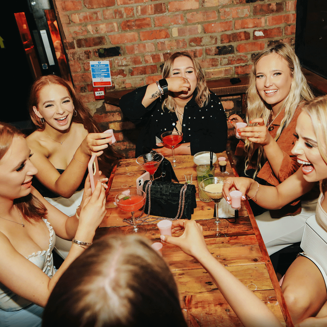 A group of six women sitting around a table in a bar, each holding drinks and laughing. They are enjoying a night out at a lively venue. The setting features a brick wall background, and colorful cocktails are on the table, emphasizing the vibrant atmosphere of Thompsons Belfast Night Club.