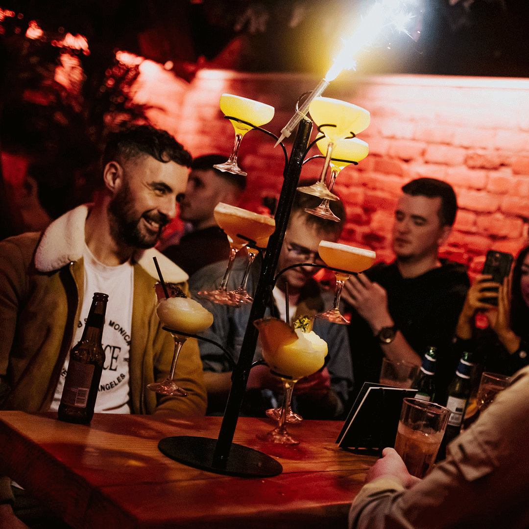 A group of people enjoying drinks at a bar, with a tall stand holding multiple yellow cocktails. The atmosphere is lively, highlighted by warm lighting reflecting off brick walls. This captures a party vibe at Thompsons Belfast Night Club.
