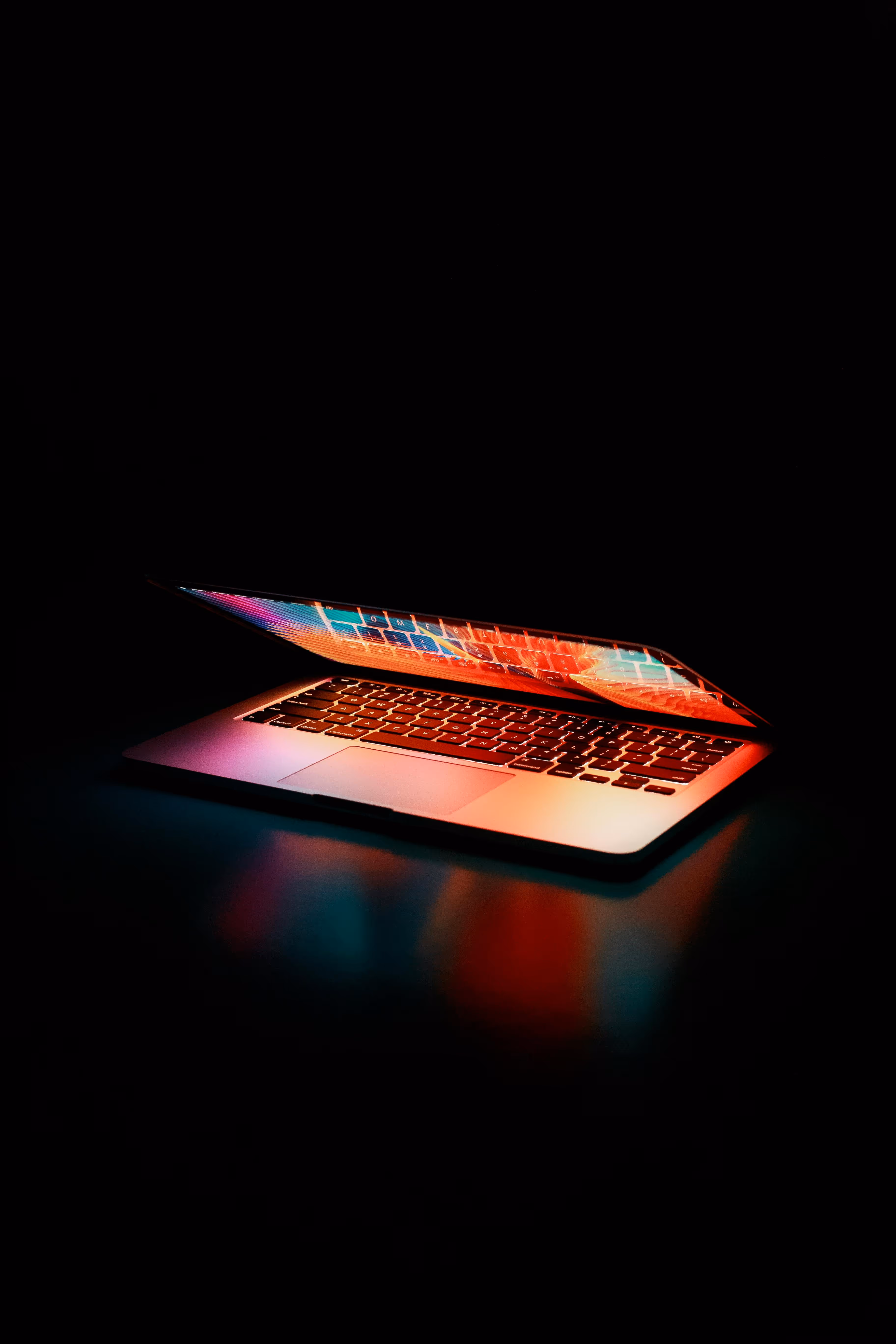 Ordinateur portable partiellement ouvert dans une pièce sombre, éclairé par des lumières colorées et reflétant des teintes vives sur le clavier et la surface, créant une ambiance futuriste et technologique.