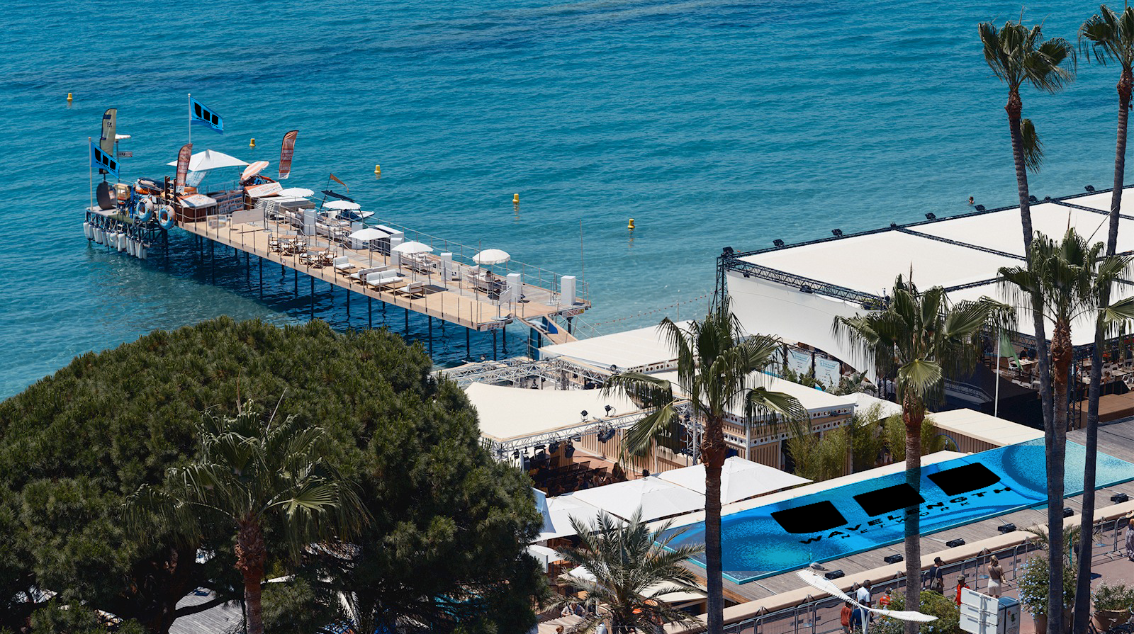Pier extending into clear blue water with lounge chairs, umbrellas, palm trees, and poolside area.