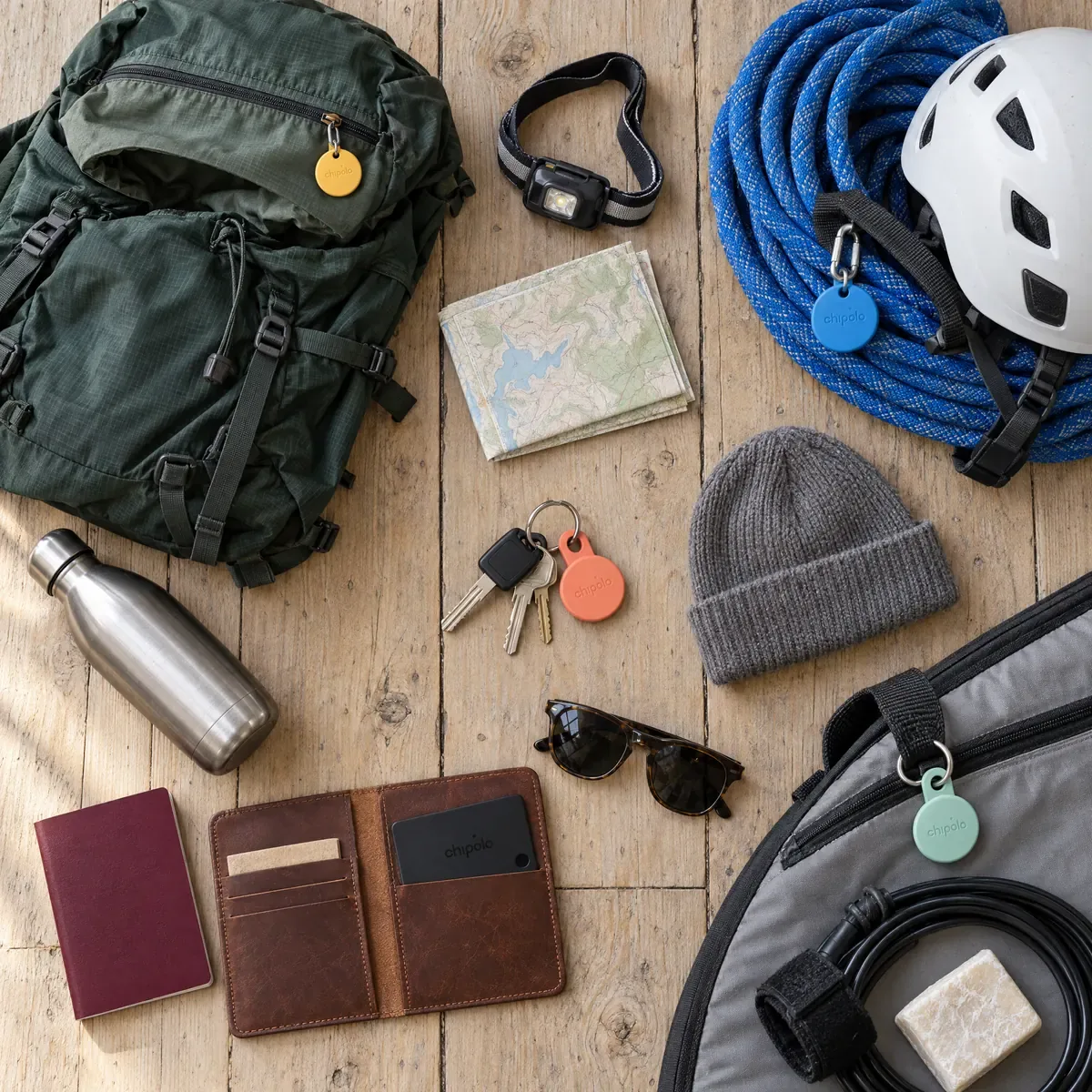 Top-down flat lay of adventure kit on a van floor: a backpack with a yellow Chipolo, a climbing rope and helmet with a blue Chipolo, van keys with a coral Chipolo LOOP, a passport wallet holding a black Chipolo CARD, and a surfboard bag with a mint Chipolo LOOP