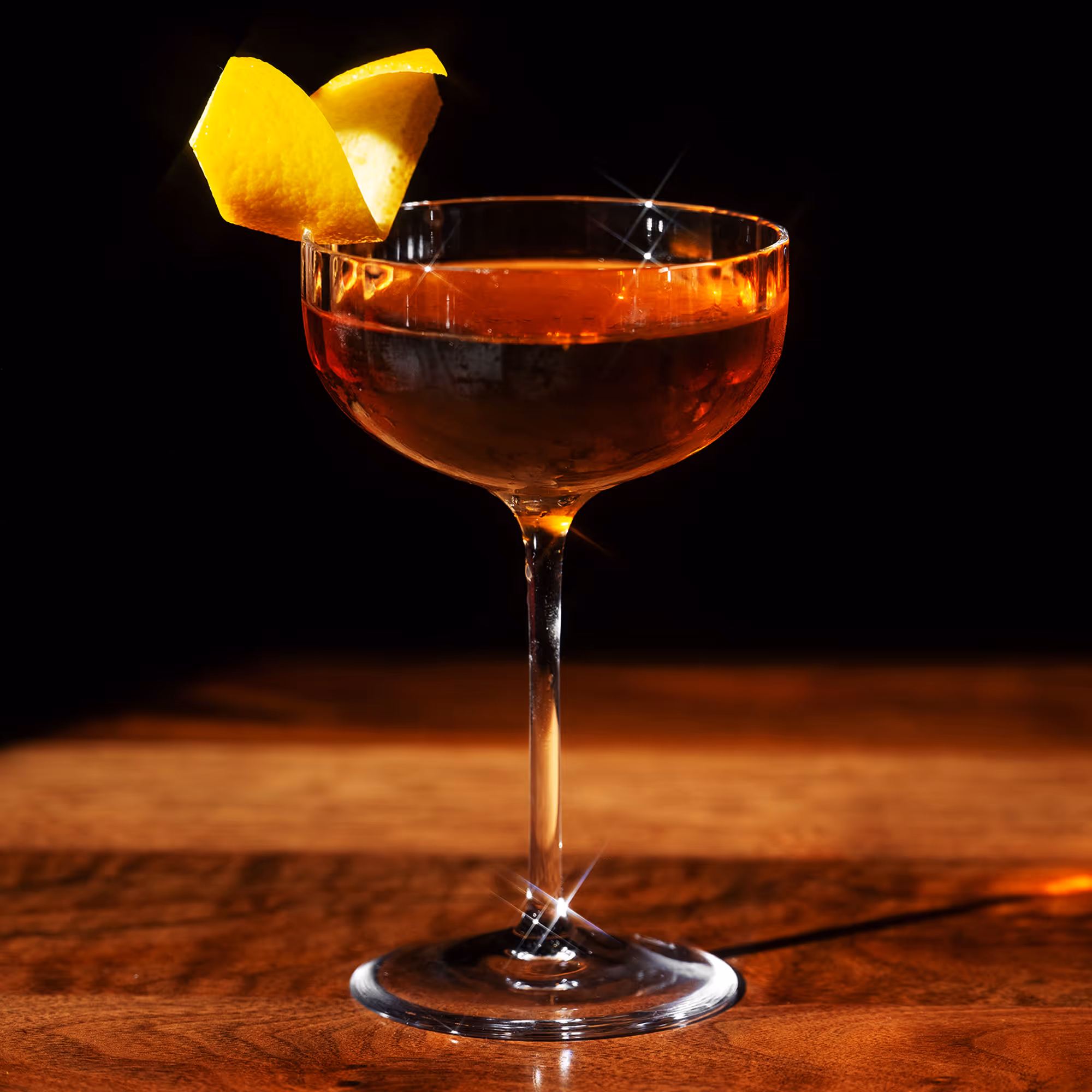 Cocktail in a coupe glass garnished with a lemon twist on a dark wood surface with black background.