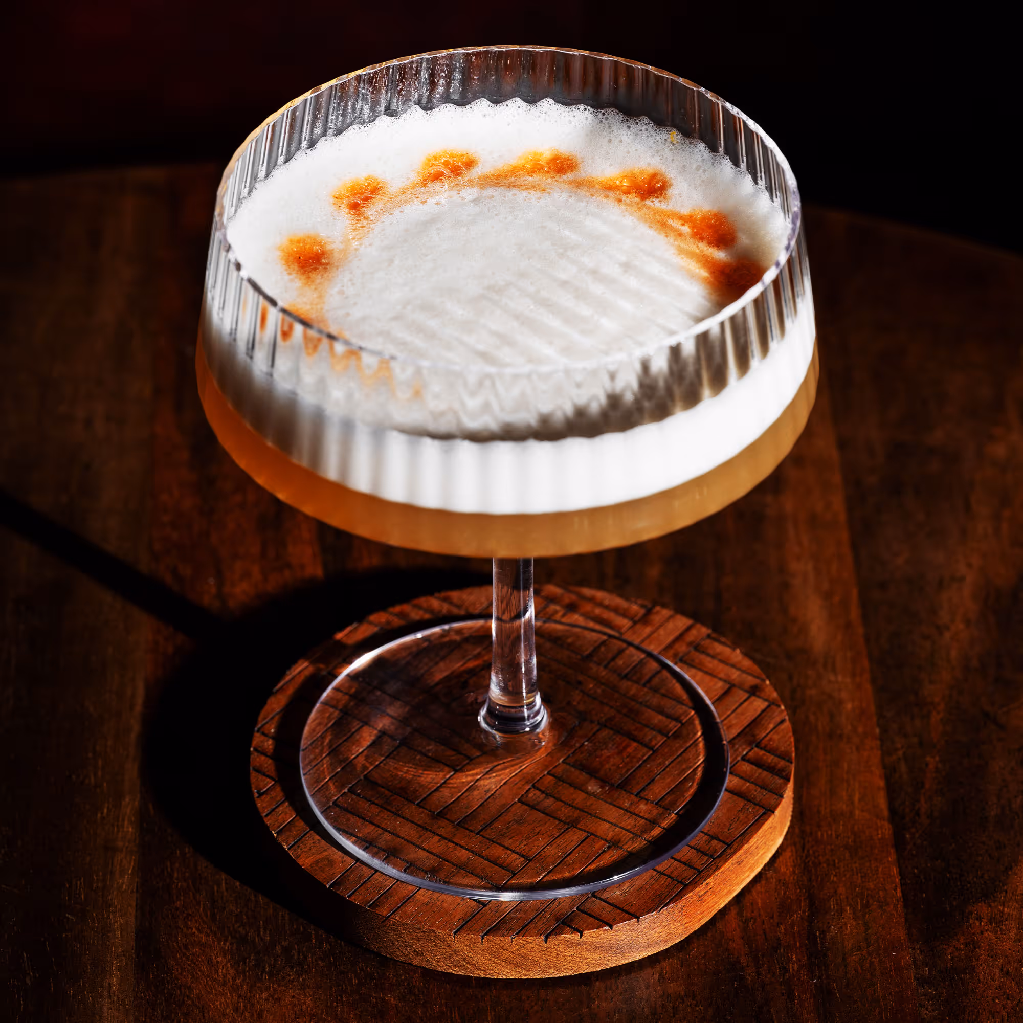 Cocktail glass with a creamy frothy top and orange bitters decoration on a wooden coaster.