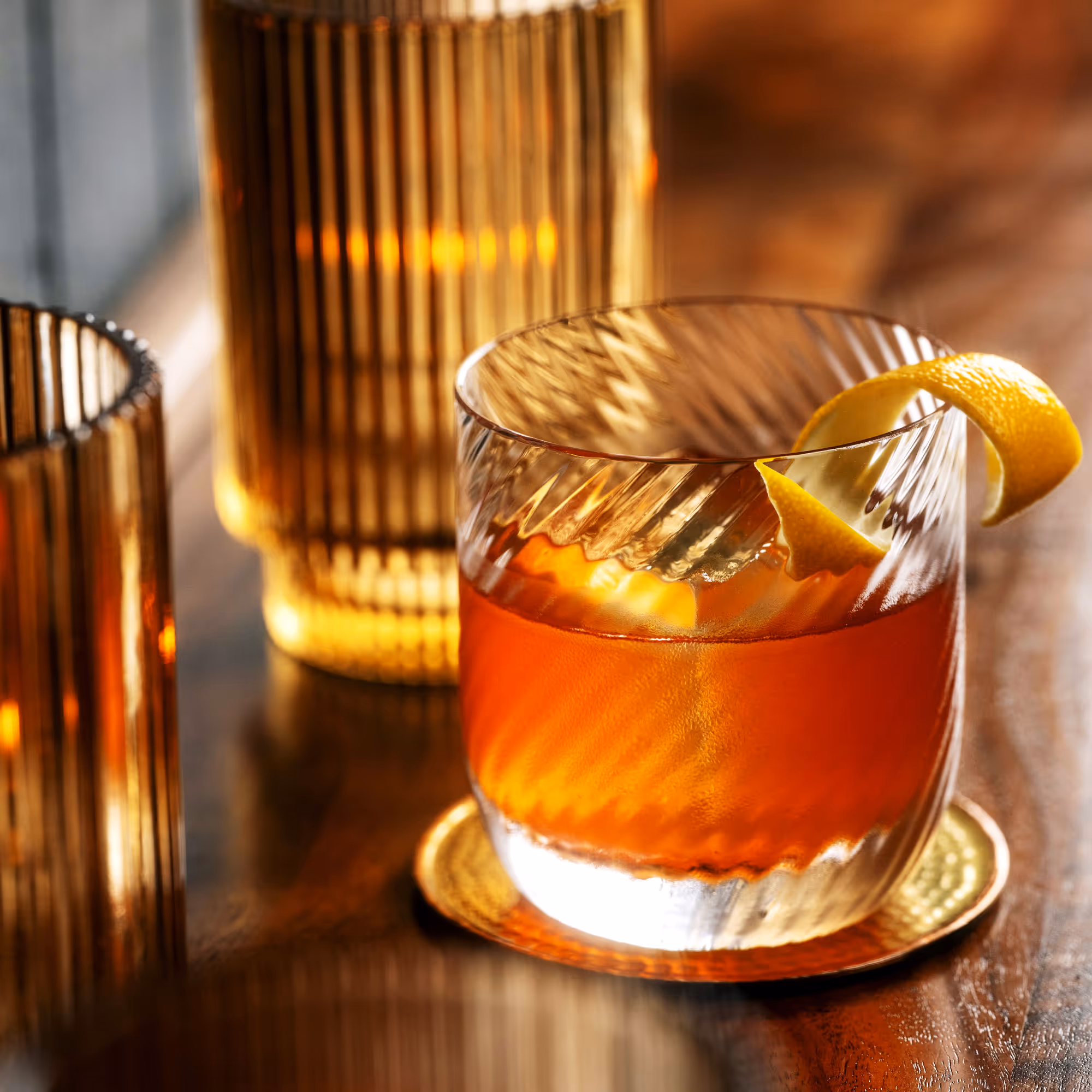 Glass of amber cocktail on a wooden surface with a lemon twist garnish and textured amber glassware in the background.
