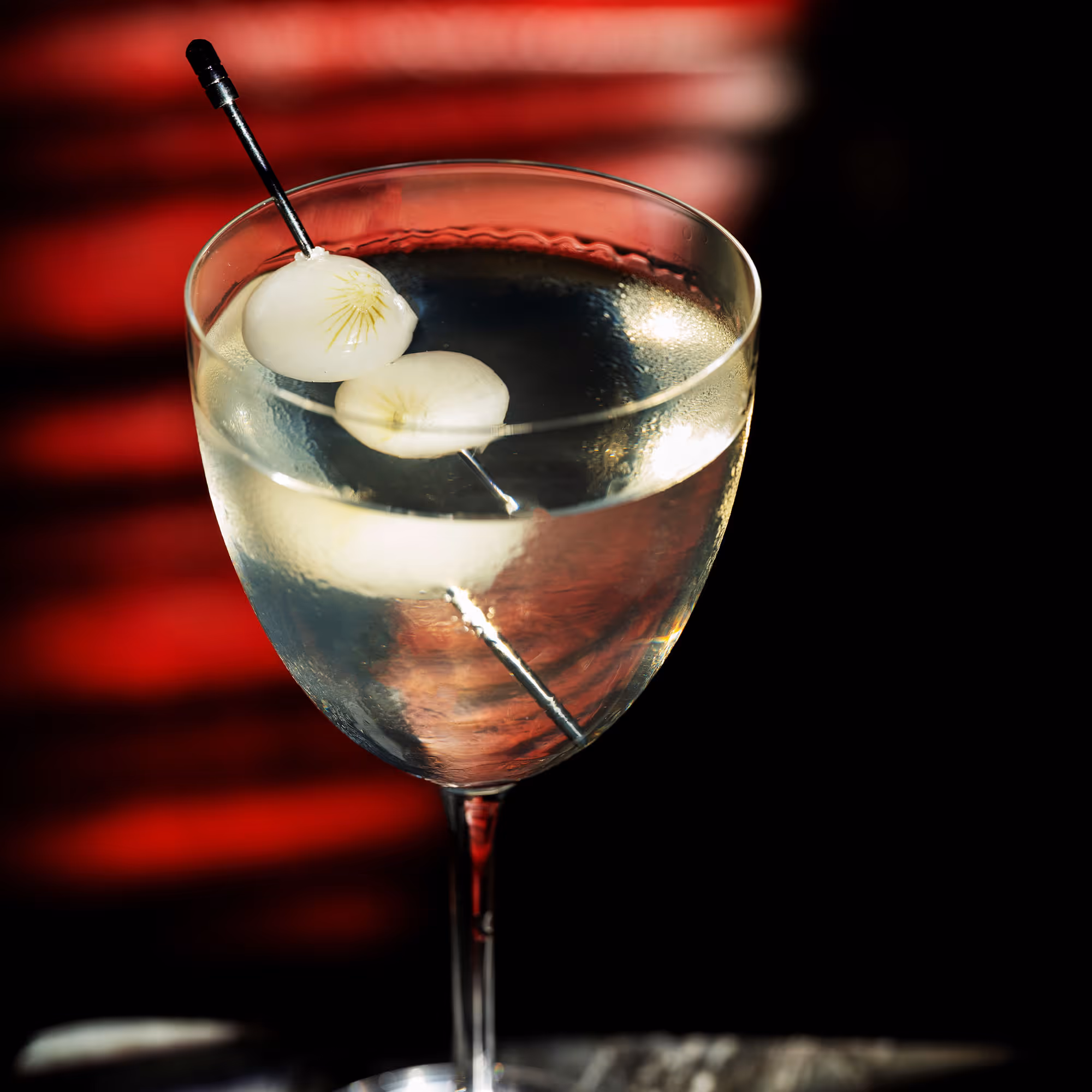 Glass of clear cocktail garnished with two white onions on a cocktail pick against a red and black background.