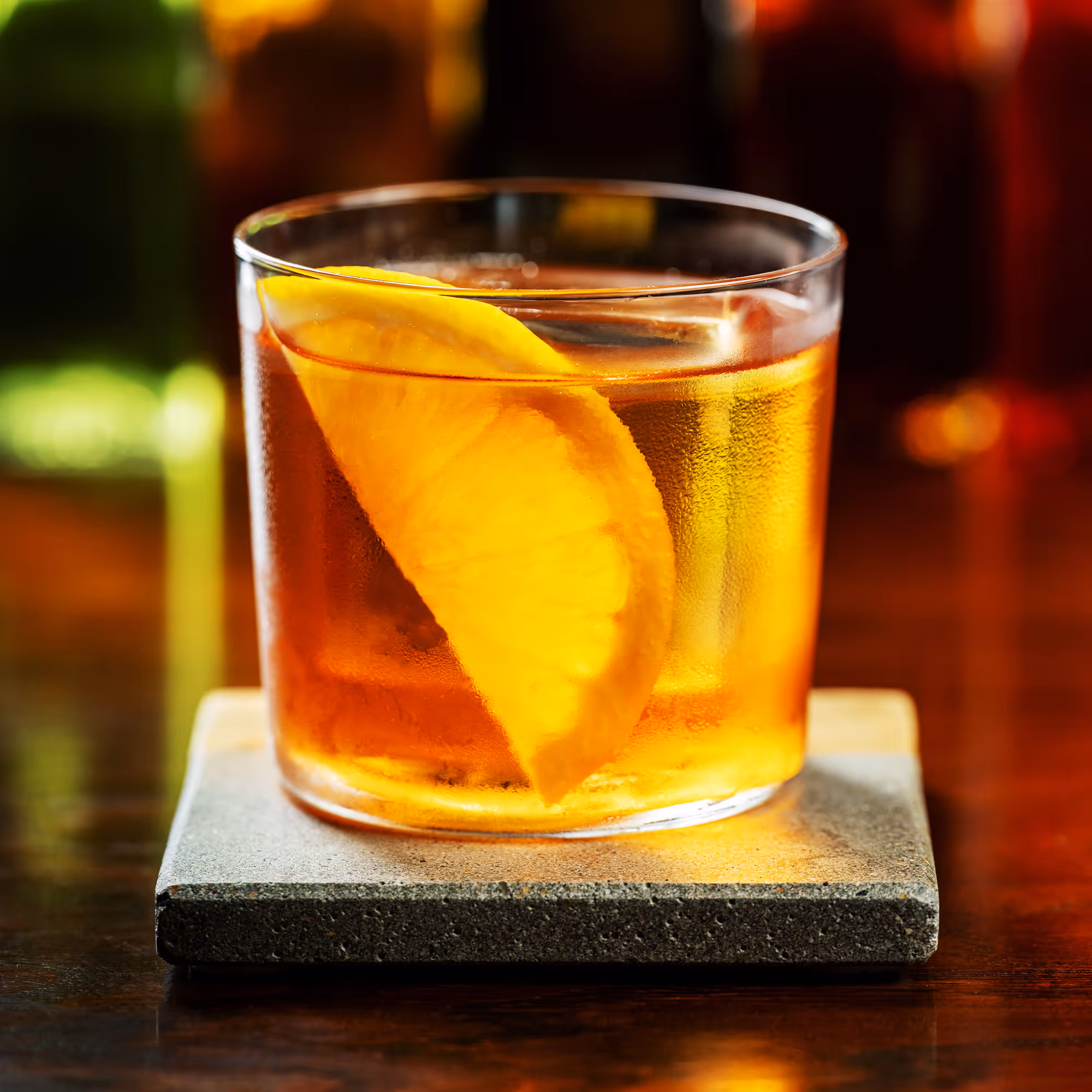 Glass of amber-colored cocktail with an orange slice on a stone coaster.