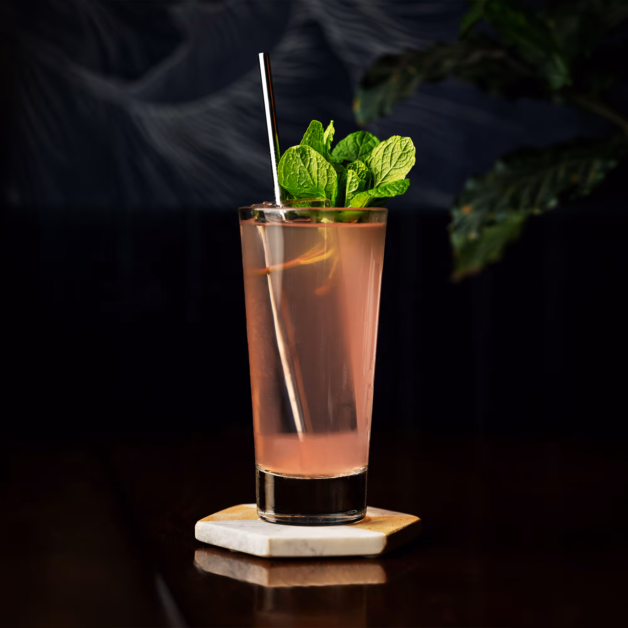 Tall glass of pink cocktail garnished with fresh mint leaves and a metal straw on a hexagonal marble coaster.