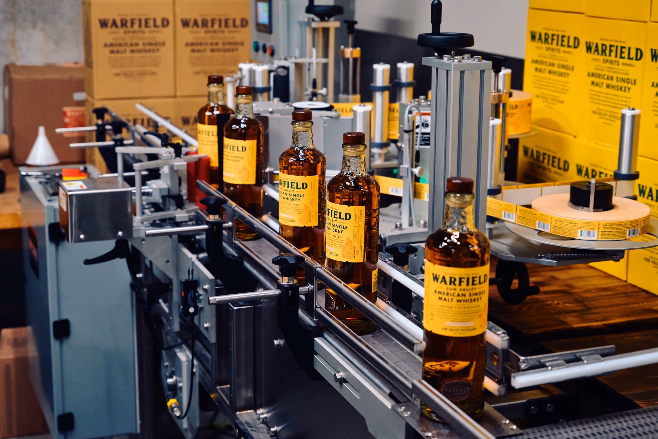 Bottles of Warfield American Single Malt Whiskey on a labeling machine conveyor in a distillery.