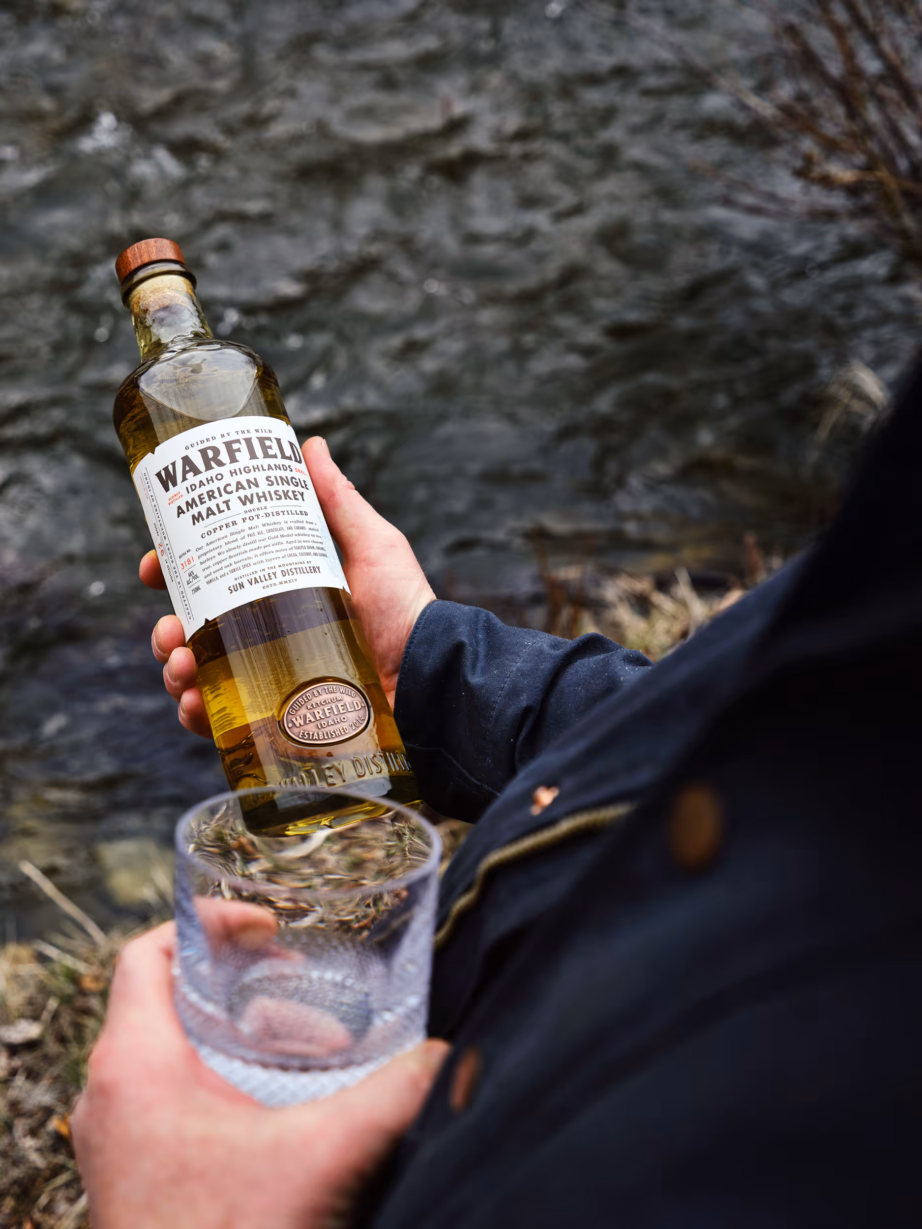 Person holding a bottle of Warfield Idaho Highlands American Single Malt Whiskey and an empty glass near a river.