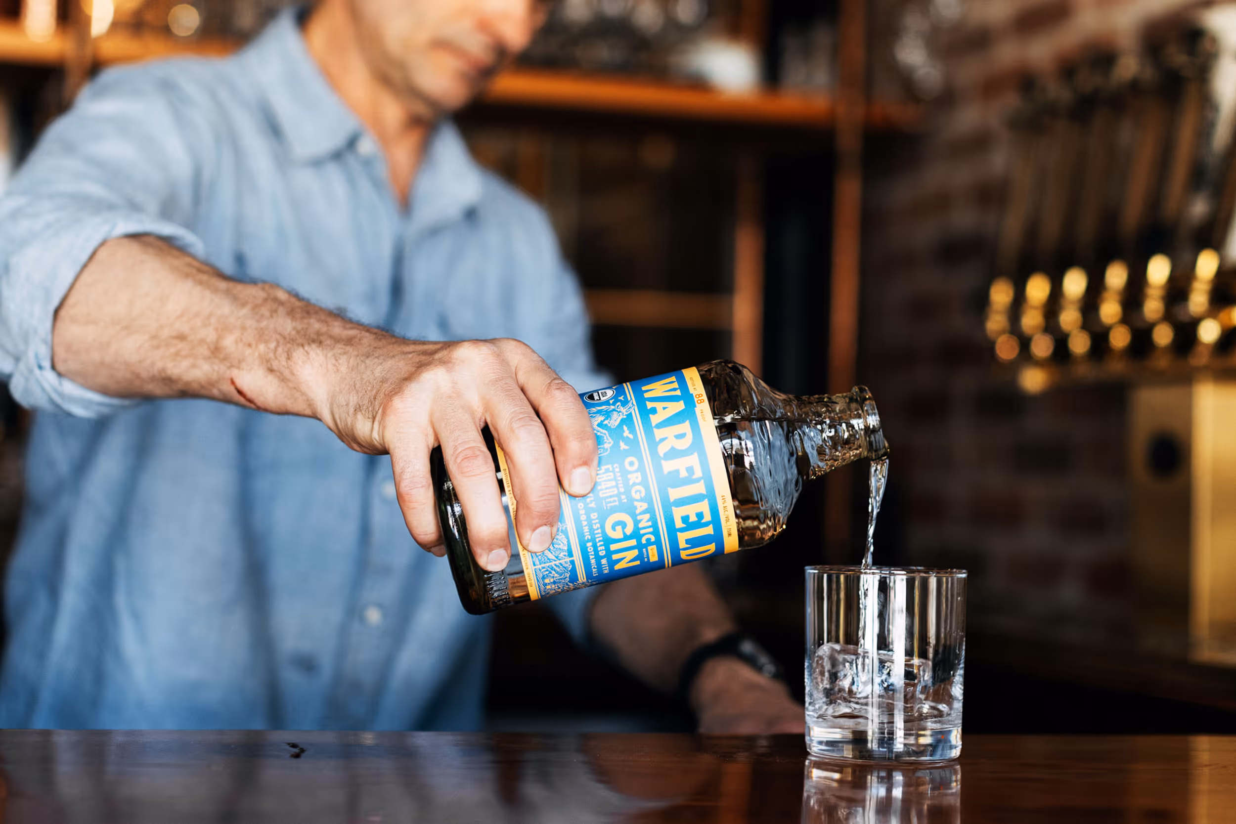 Person pouring Warfield Organic Gin from a bottle into a glass with ice on a wooden bar counter.