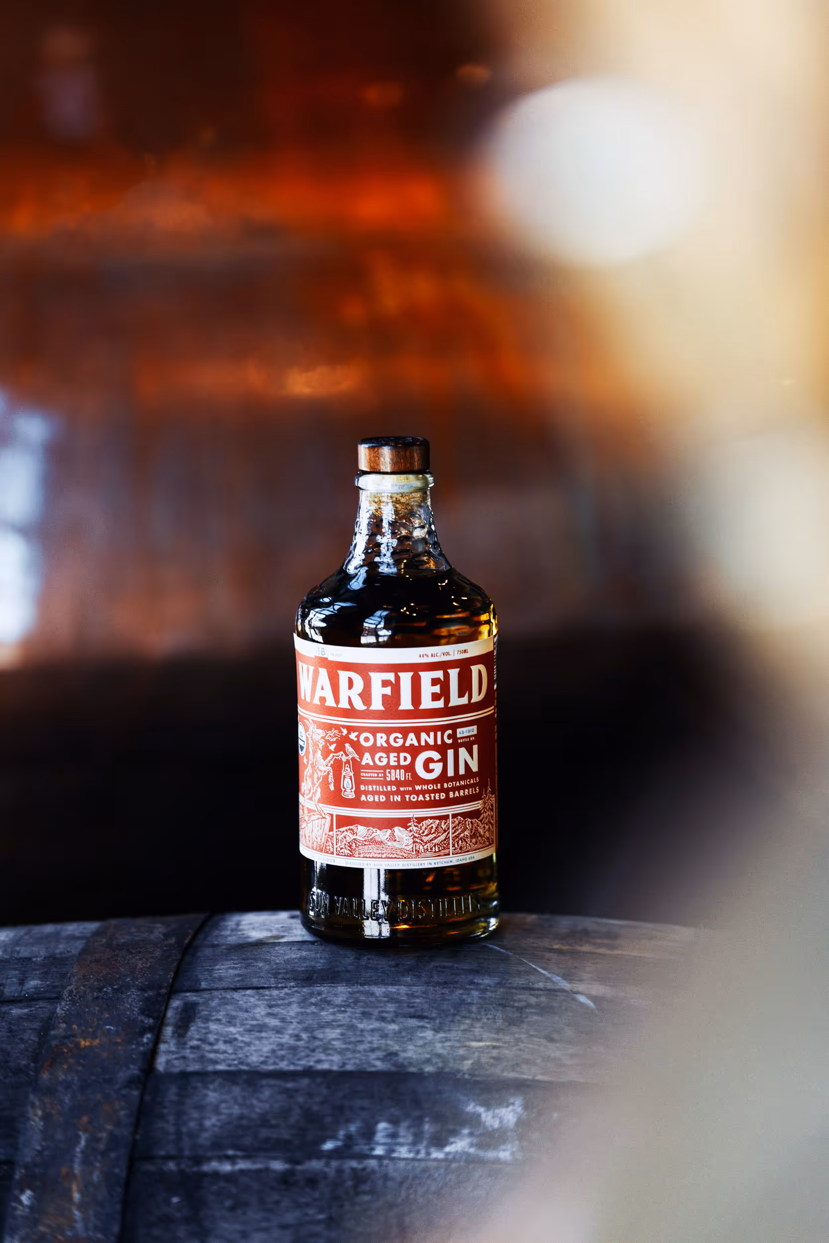 Bottle of Warfield Organic Aged Gin with a red label sitting on a wooden barrel.