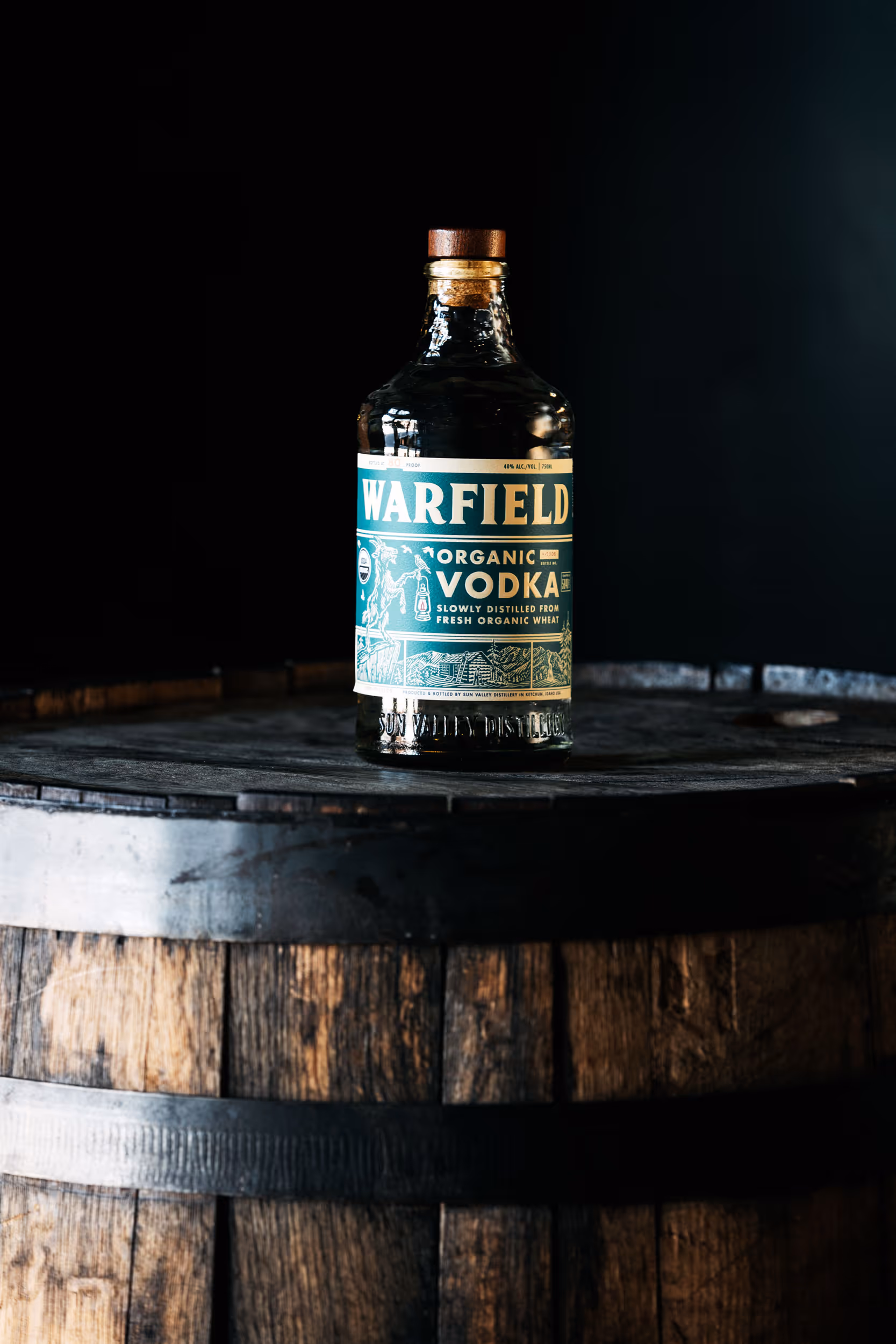 Bottle of Warfield organic vodka placed on top of a wooden barrel against a dark background.