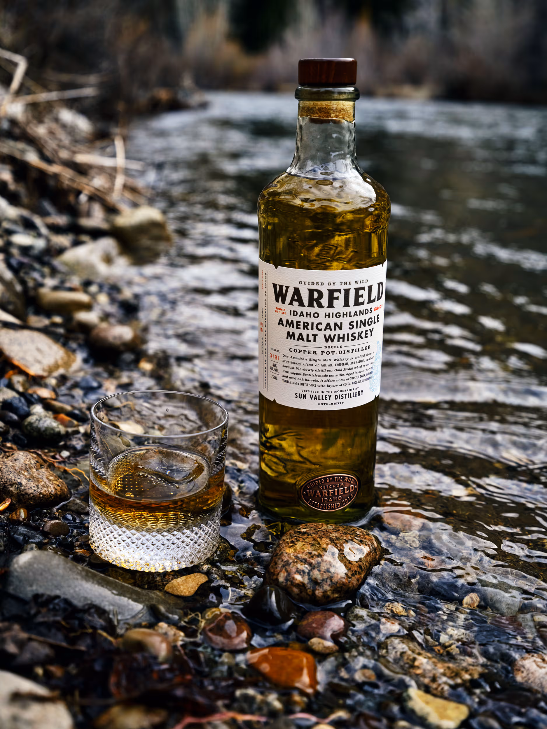 A bottle of Warfield American Single Malt Whiskey and a glass with whiskey and an ice cube on a rocky riverbank.