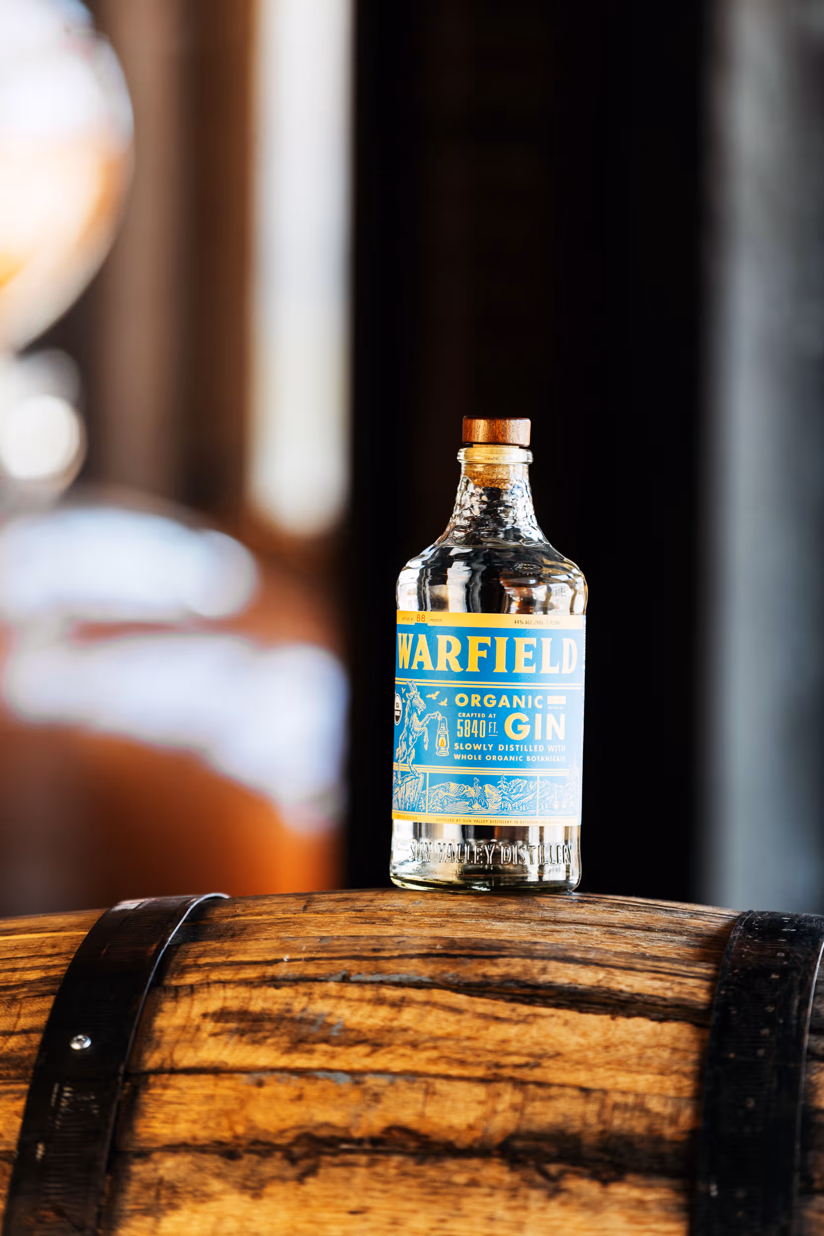 Clear glass bottle of Warfield Organic Gin with a cork stopper on a wooden barrel.