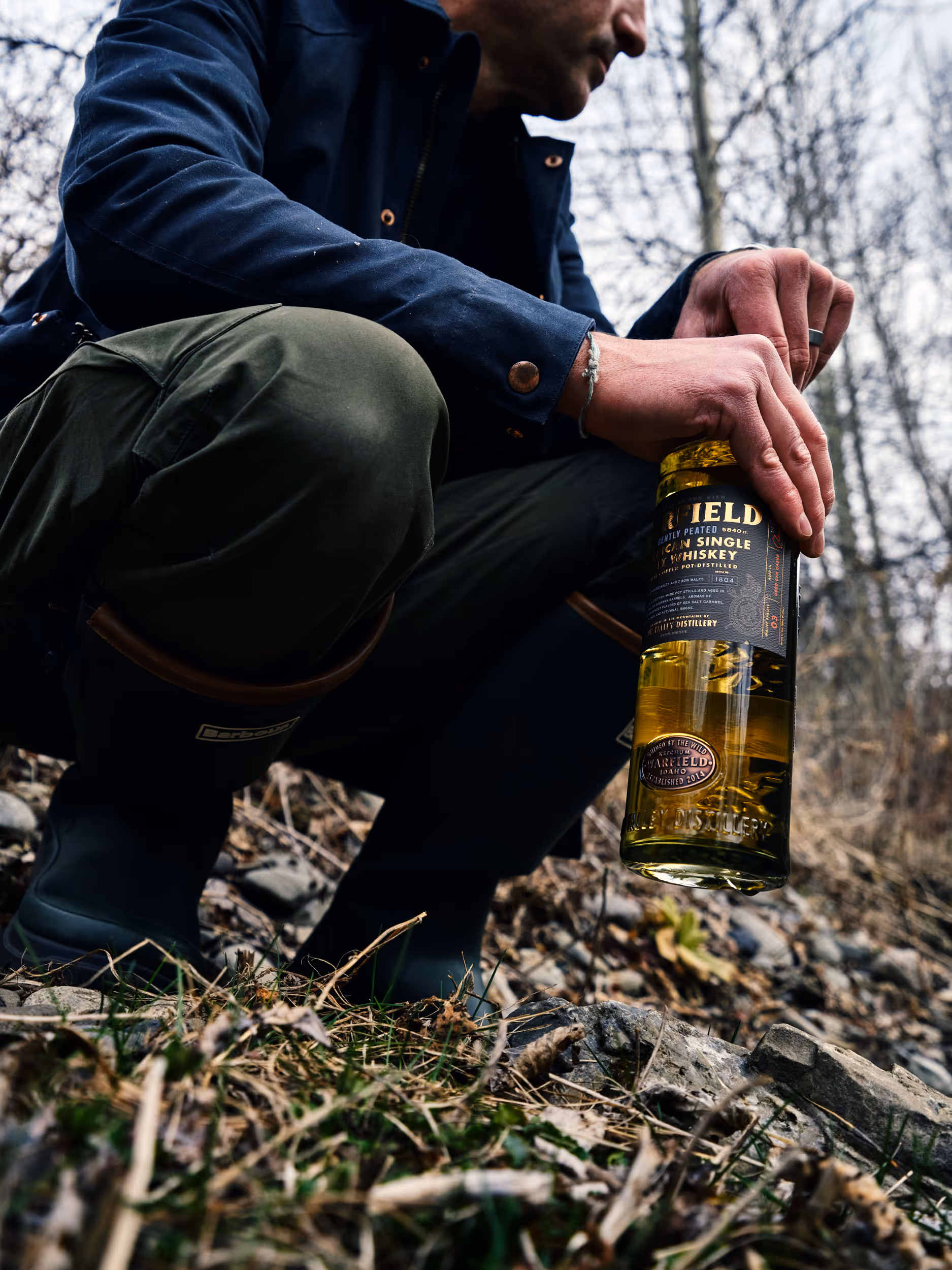 Person crouching outdoors in a wooded area holding a bottle of Warfield American Single Malt Whiskey.