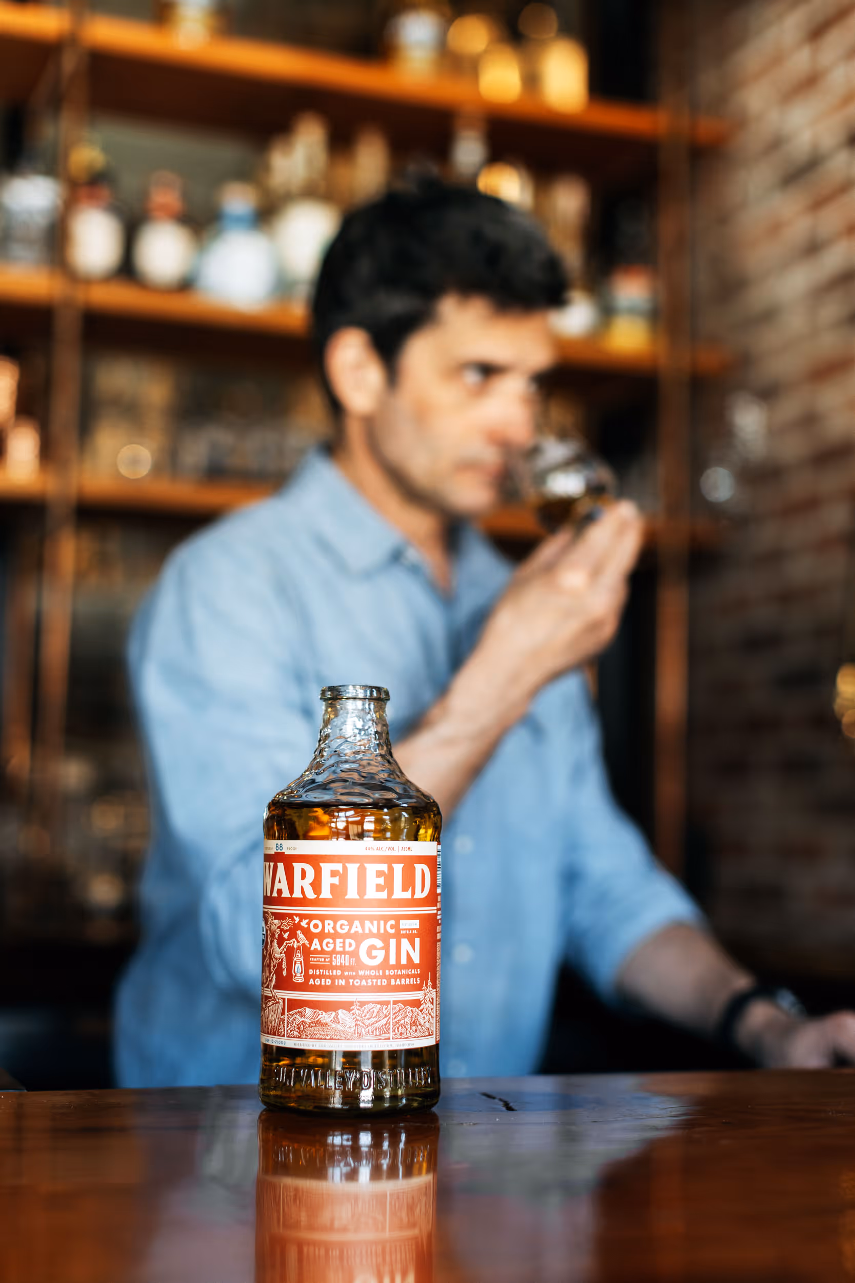 Glass bottle of Warfield Organic Aged Gin on a wooden bar with a man in the background smelling a glass of liquor.