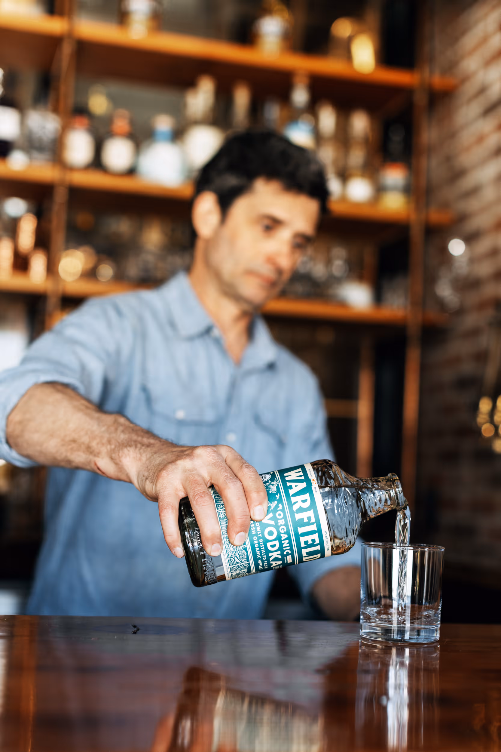 Man in a blue shirt pouring Warfield organic vodka into a clear glass at a bar.